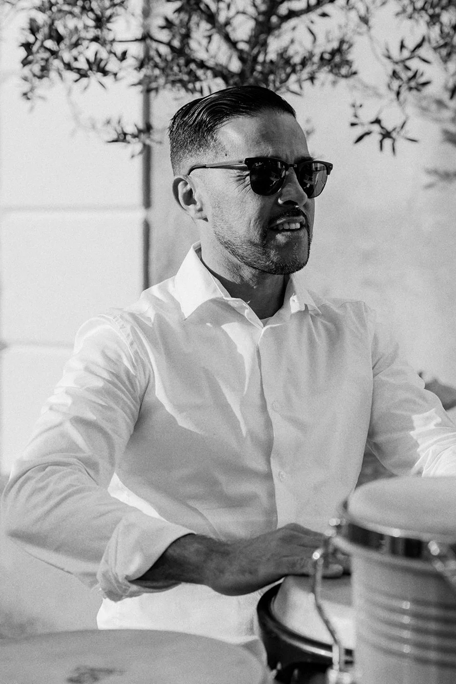 Man wearing sunglasses and a white dress shirt, playing drums outdoors in a sunny setting.