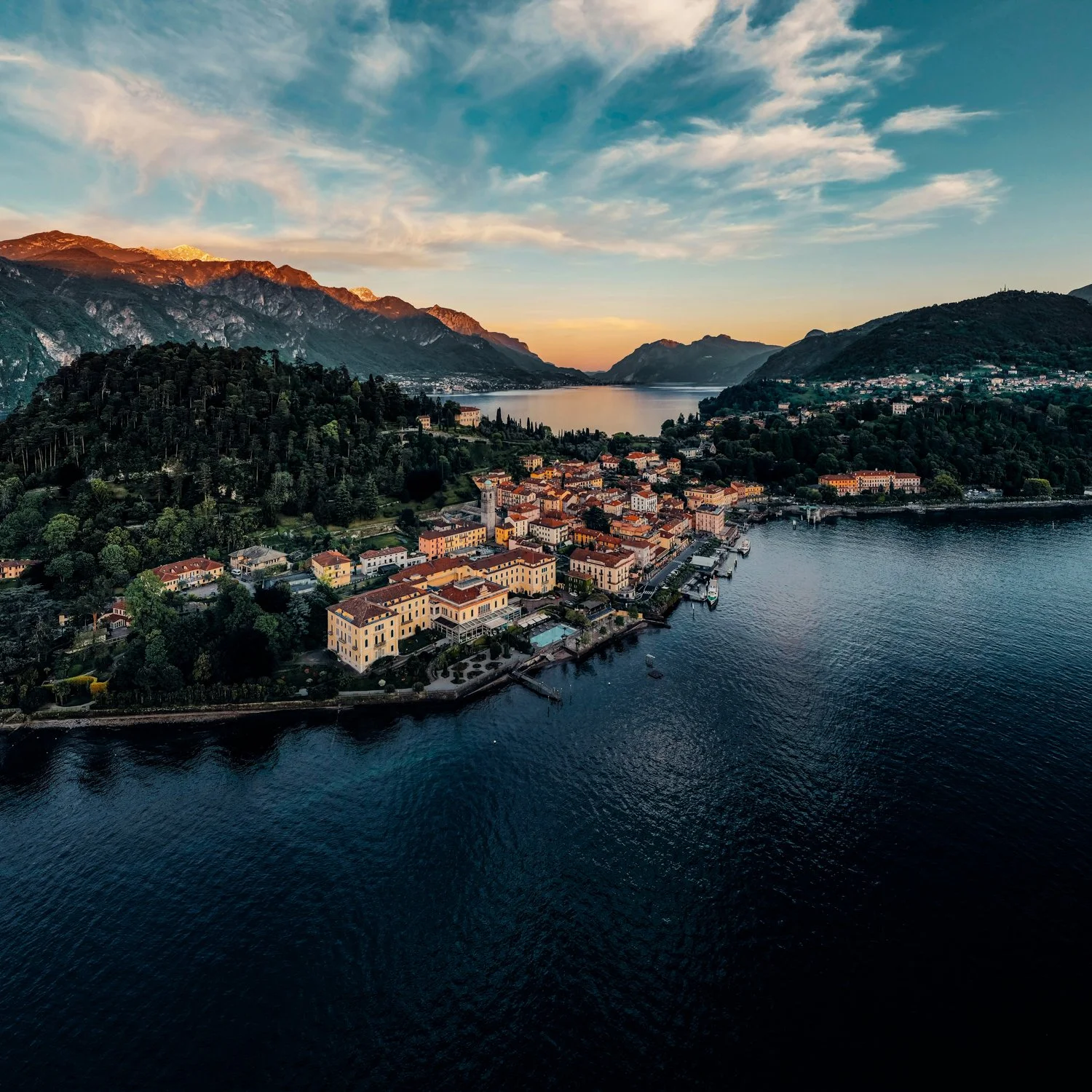 Aerial view of a lakeside town nestled among green mountains at sunset, with colorful buildings along the waterfront and boats in the water.