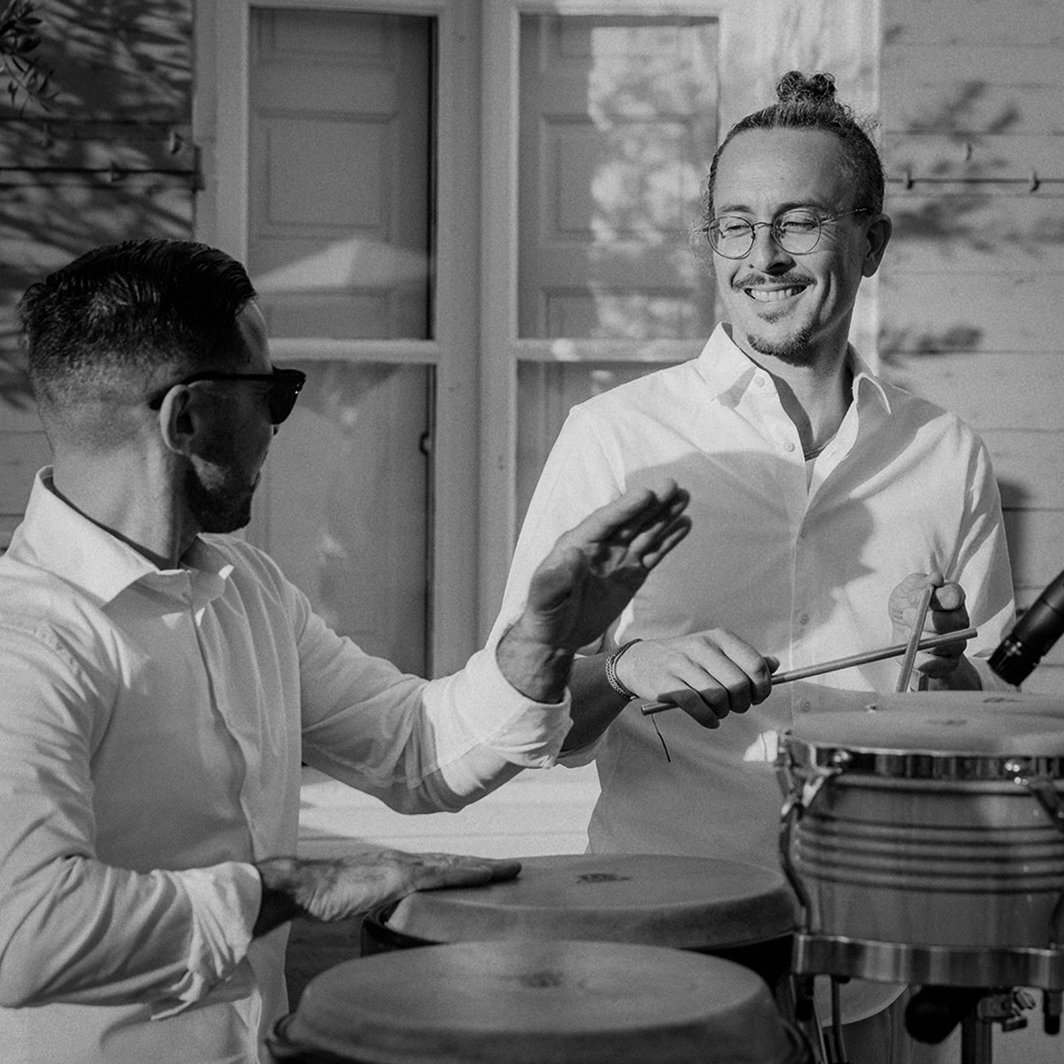 Two men playing percussion instruments indoors, smiling, one holding drumsticks, the other playing conga drums, in a bright room with windows.