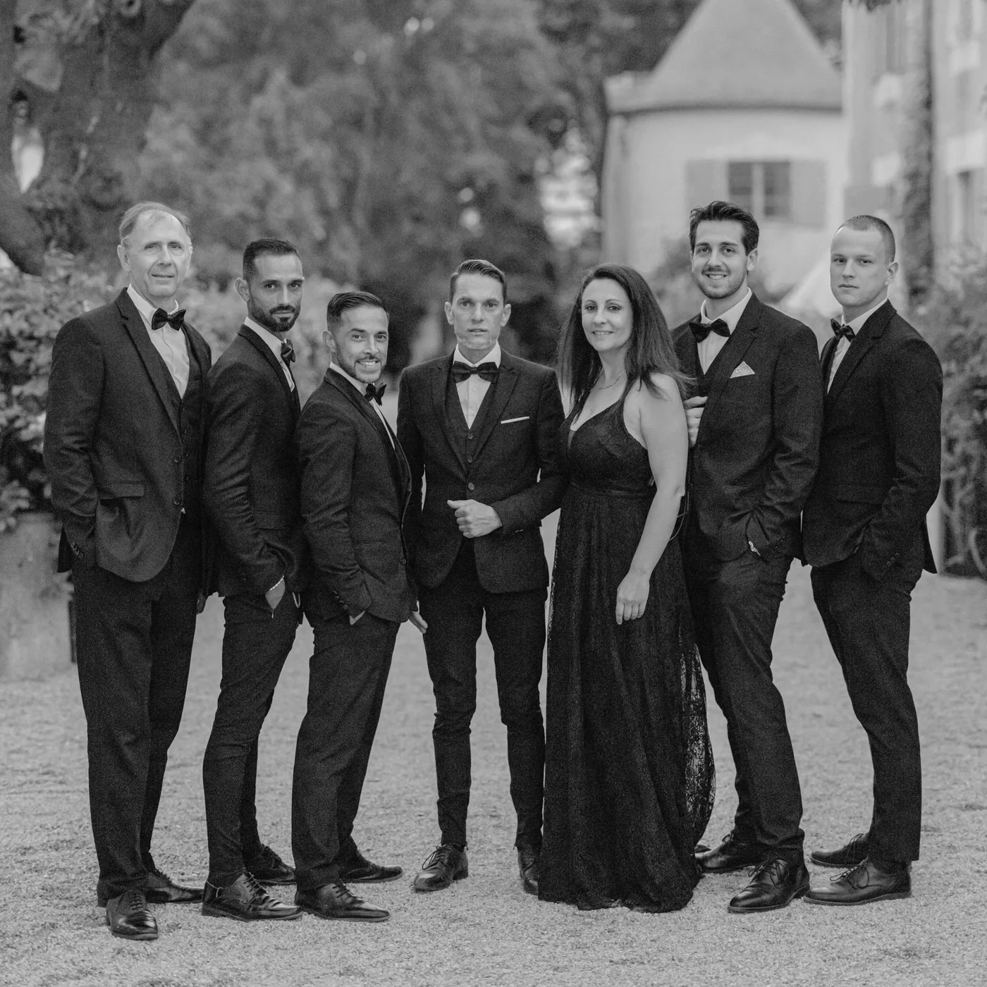 Black and white photo of eight people in formal wear standing outdoors, with trees and a house in the background.