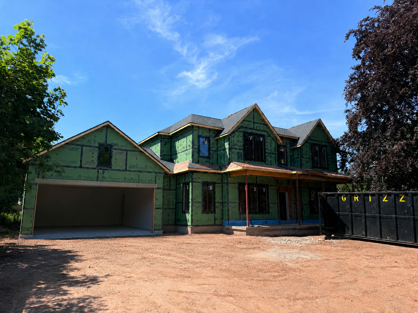 A two-story house under construction with green sheathing on the exterior walls, black window frames, and a gray shingle roof. The house is surrounded by trees and a dirt lot, with a black dumpster labeled 'GRIZZ' on the right side.