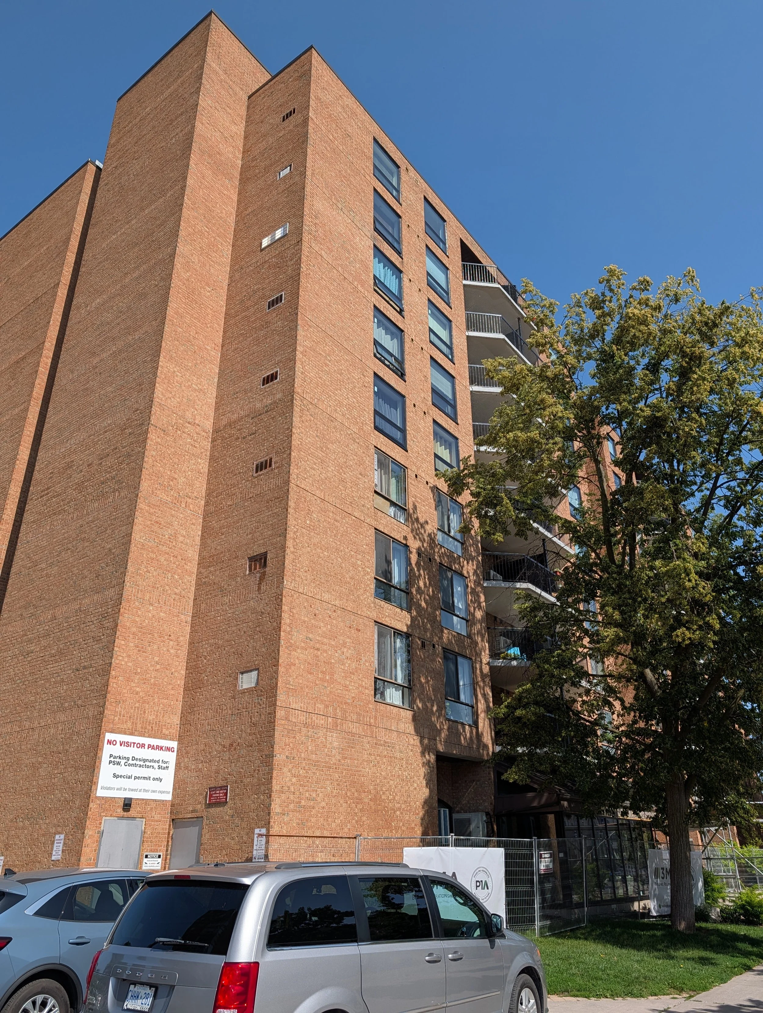 A multi-story brick residential building with several windows, including balconies on the right side. The foreground shows a silver minivan parked on the street and a tree with green leaves. There are signs on the building indicating parking restrictions and notices, and the sky is clear and blue.