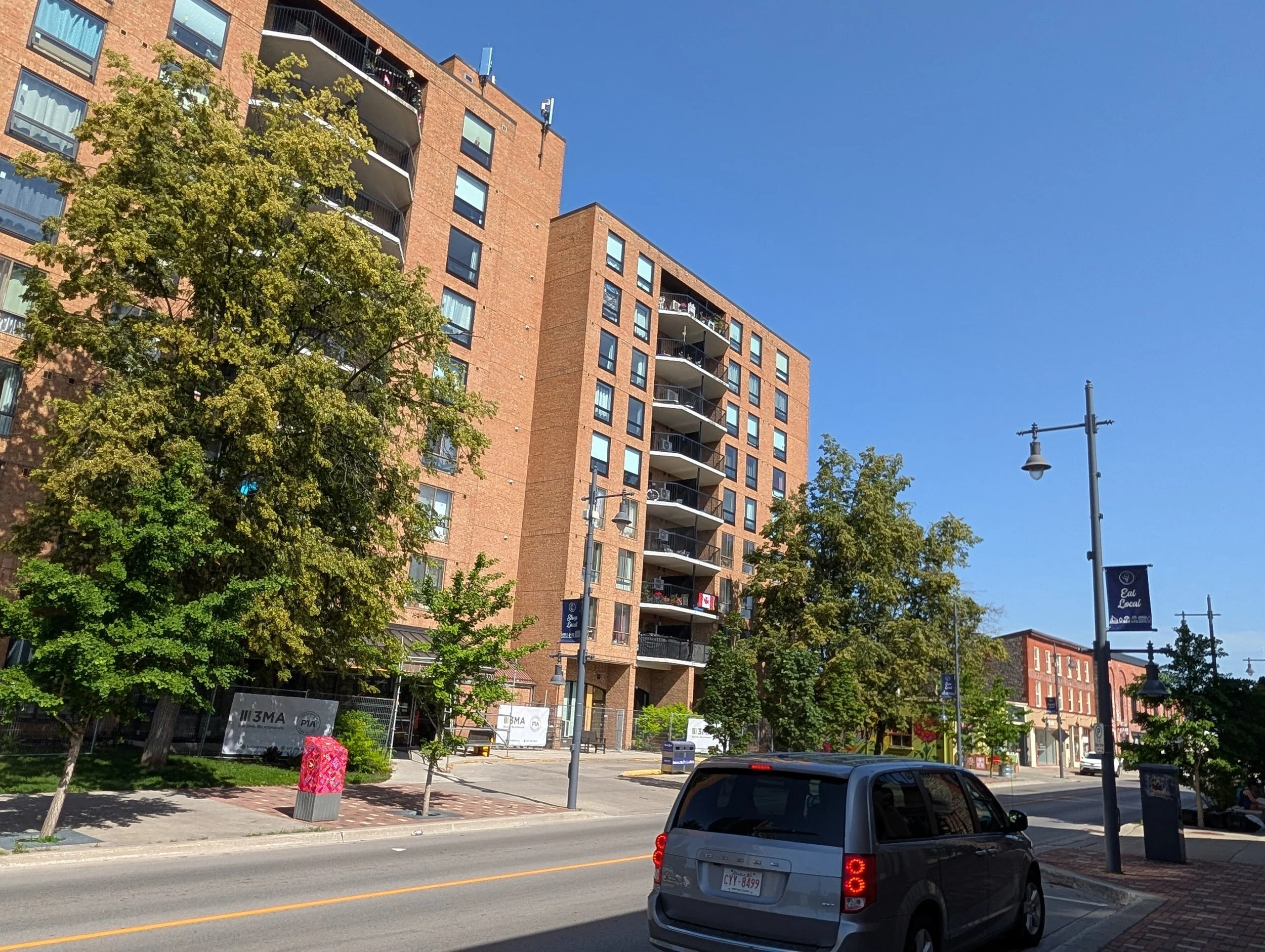 A city street scene with a high-rise brick building, trees, street lamp, and cars parked along the road on a clear day.
