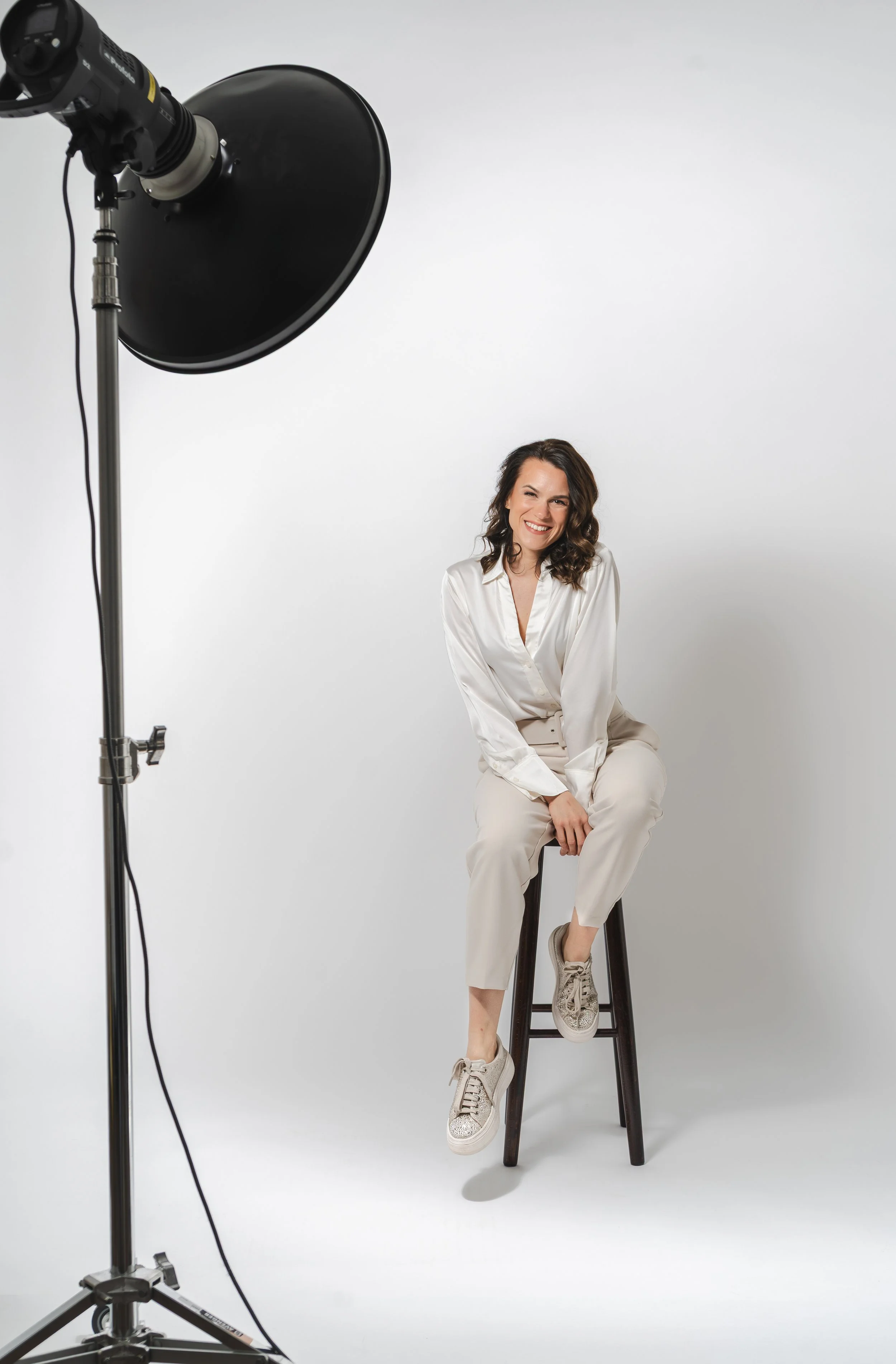Eine lachende Frau in weißen Kleidung sitzt auf einem Hocker vor einem weißen Hintergrund, während sie von einer Fotostudio-Softbox beleuchtet wird.