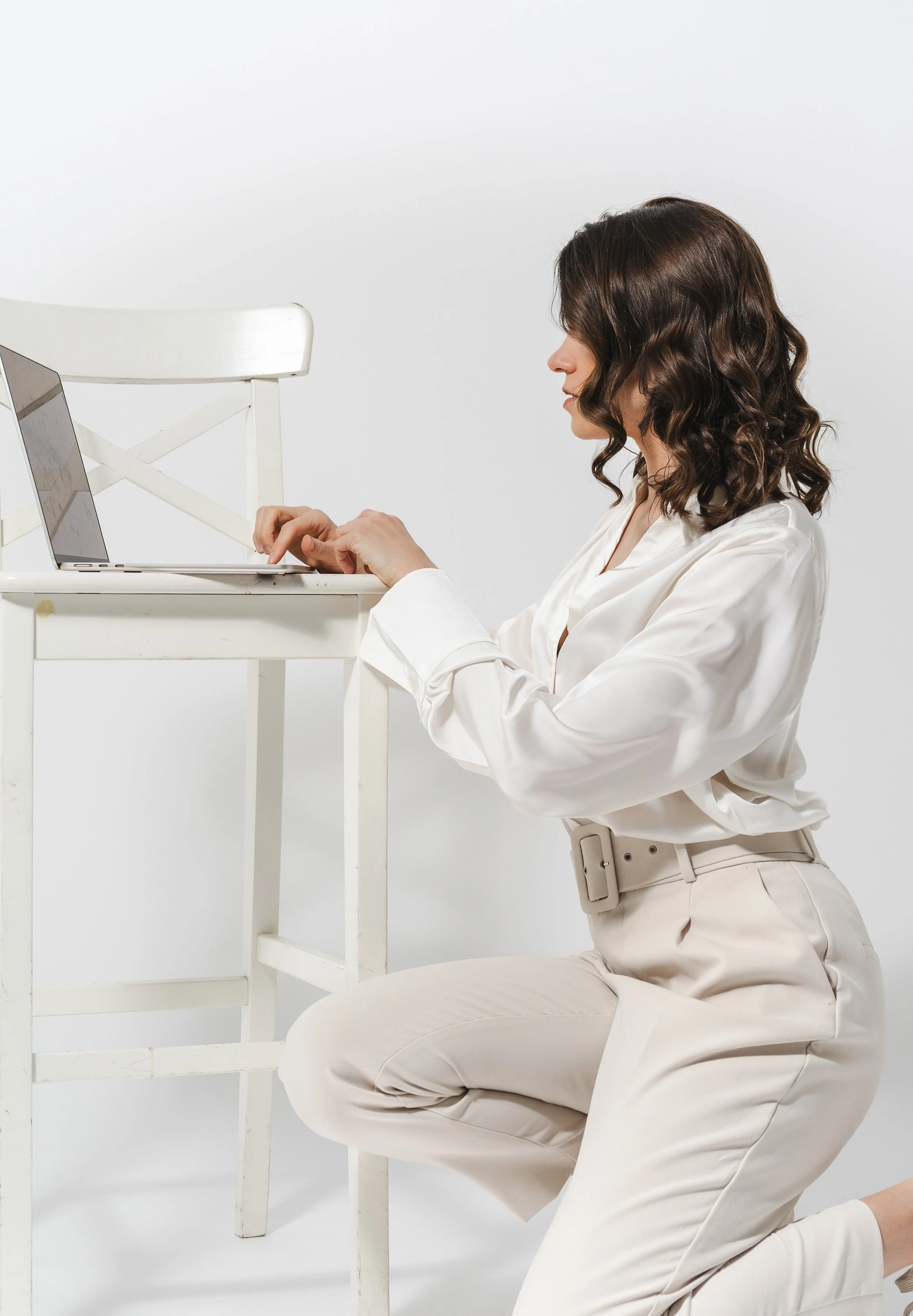 Frau in weißen Klamotten sitzt auf dem Boden, arbeitet am Laptop auf einem weißen Tisch, hat braune, lockige Haare, vor einem weißen Hintergrund.