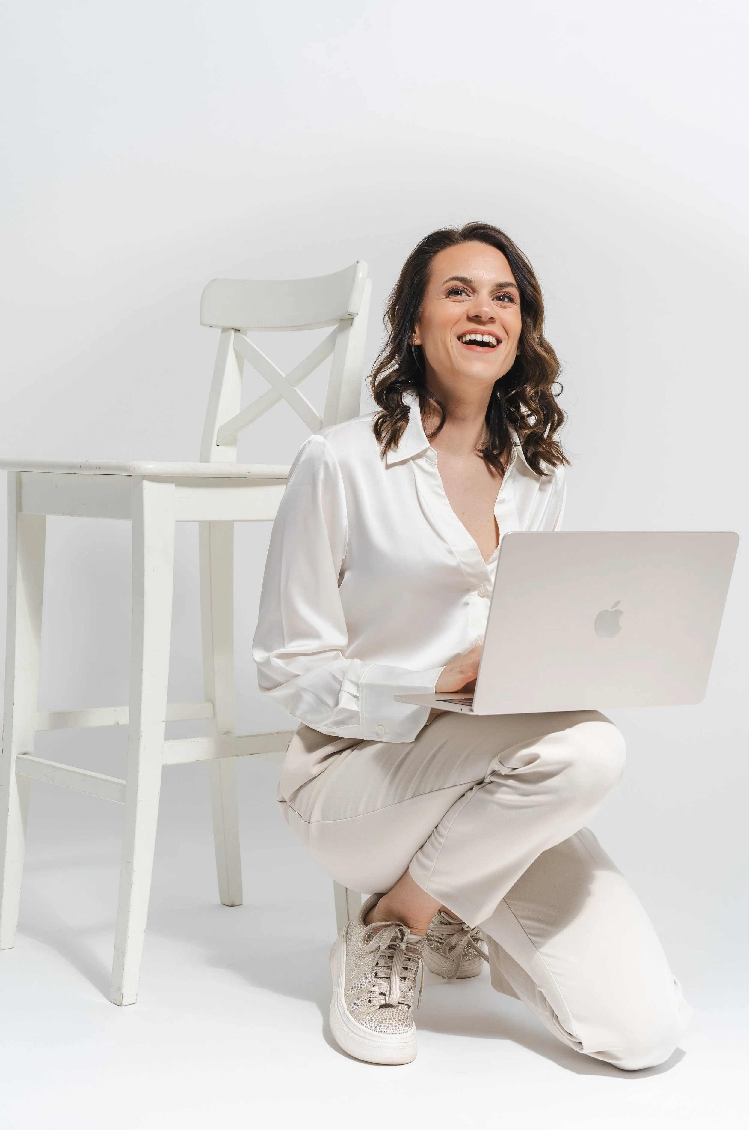 Eine lachende Frau sitzt auf dem Boden vor einem Laptop, während sie in Weiß gekleidet ist, mit einer weißen Stuhl im Hintergrund und einem weißen Hintergrund.