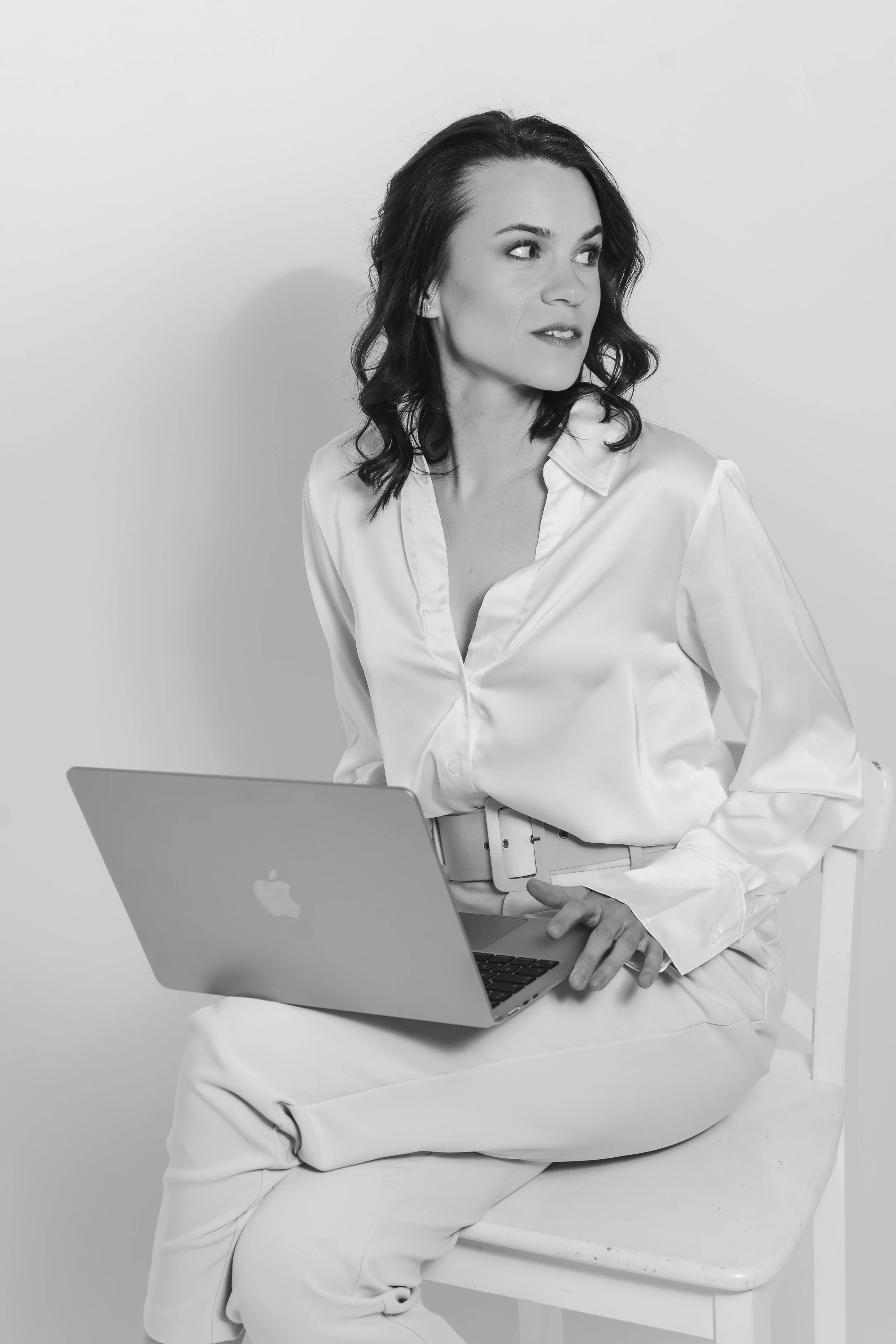 Schwarz-weiß-Foto einer Frau mit dunklen, gewellten Haaren, die in Weiß gekleidet ist, während sie auf einem Stuhl sitzt und an einem Laptop arbeitet.