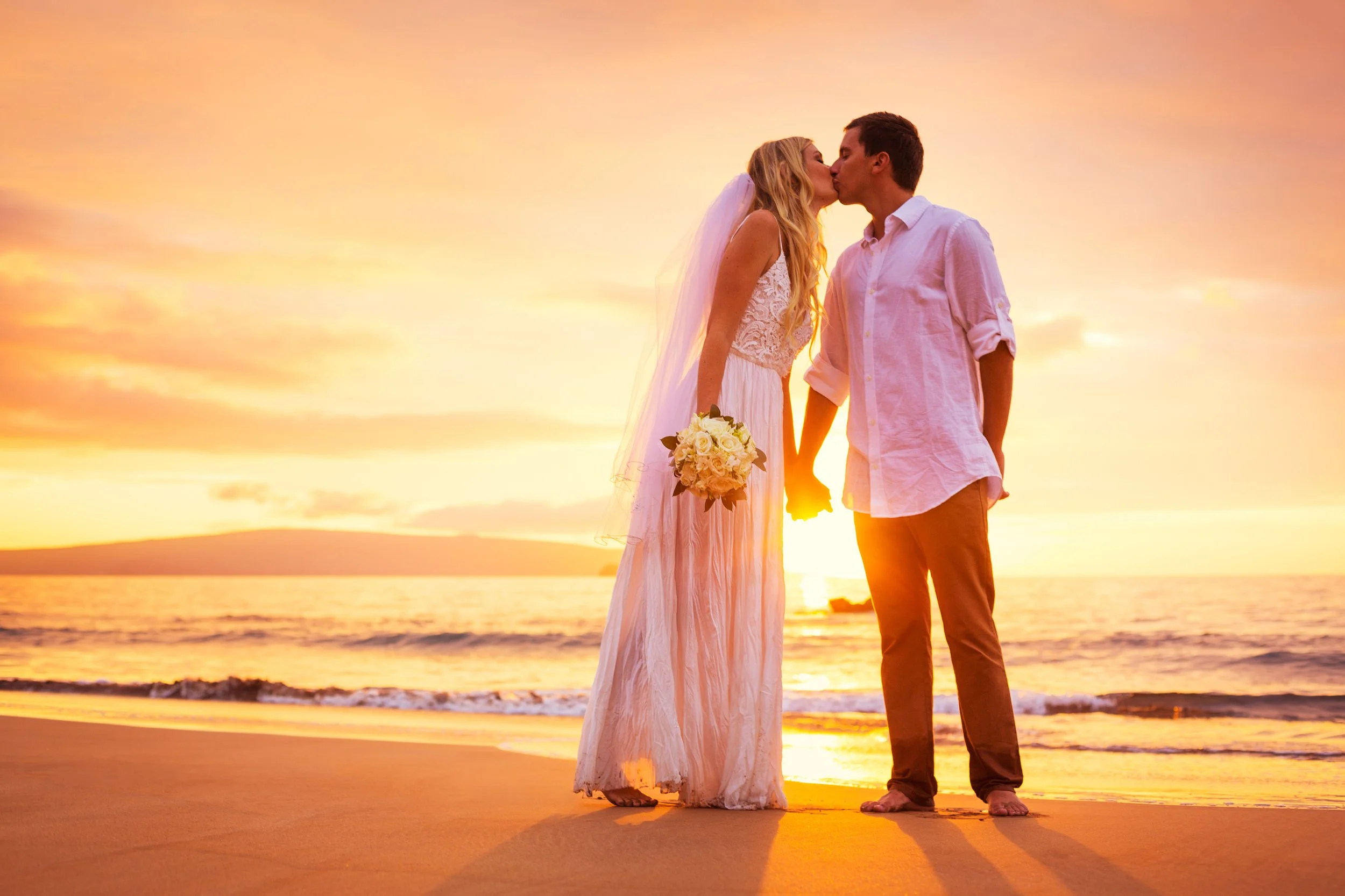 A couple sharing a kiss on the beach at sunset, the bride holding a bouquet of flowers and the couple holding hands.