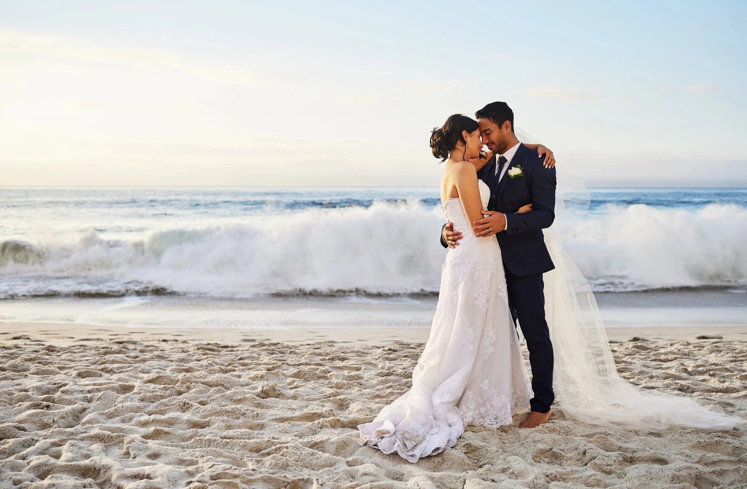 A bride and groom hugging and touching foreheads on the beach during their wedding, with waves and the ocean in the background.