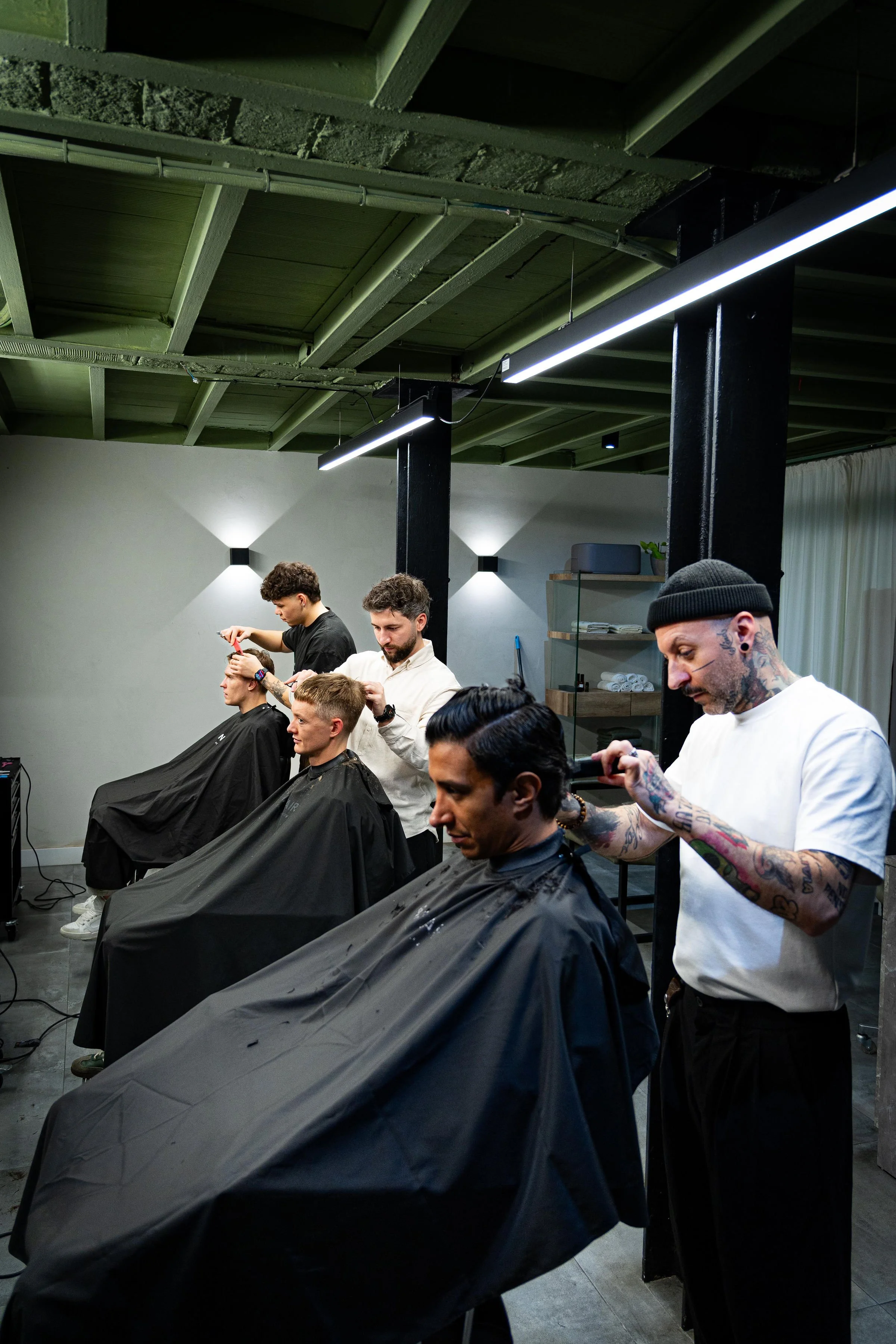 Barbers giving haircuts to clients in a modern barbershop, with stylists standing behind clients, using scissors and clippers, black capes draped over clients, and the shop has a contemporary industrial design with exposed ceiling beams and stylish lighting.