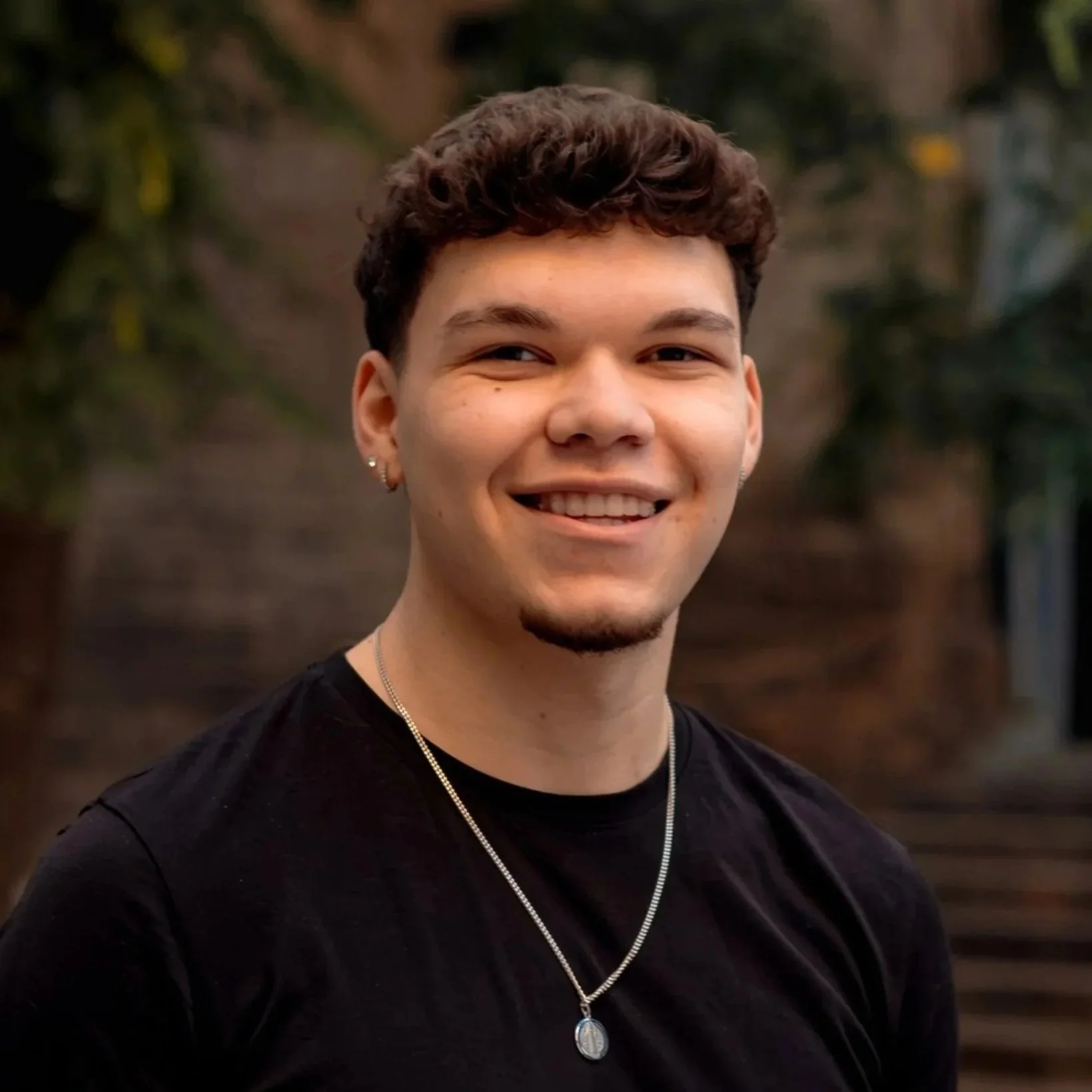 A young man with short curly hair, earrings, and a goatee smiling outdoors, wearing a black shirt and a silver necklace with an oval pendant, with blurred trees and stairs in the background.