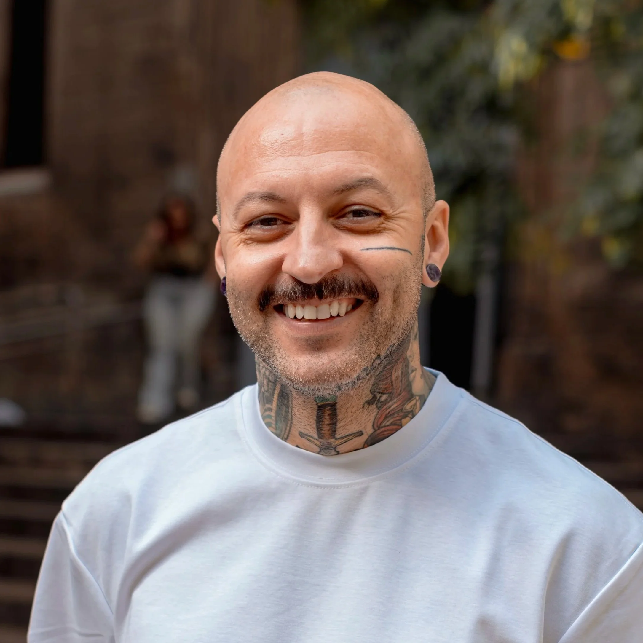 Close-up of smiling, bald man with tattoos and earrings, wearing a white t-shirt, outdoors in front of brick stairs and a woman in the background.