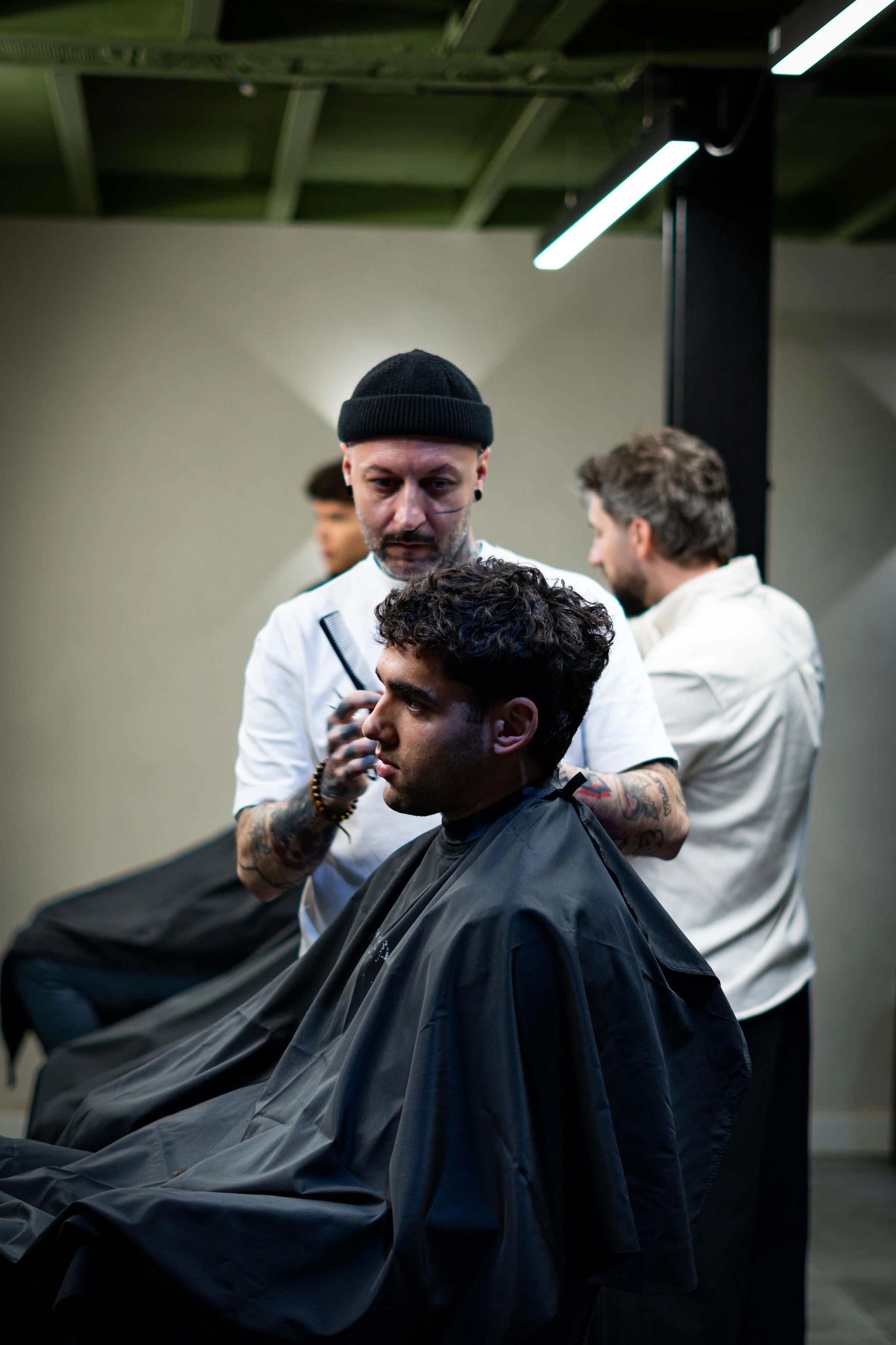 Barber cutting a young man's hair in a salon with two other people in the background.
