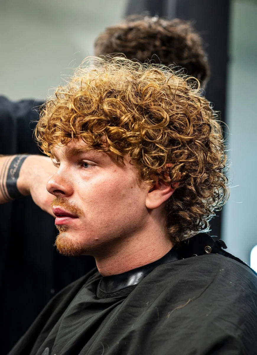 A young man with curly blond hair getting a haircut at a salon.