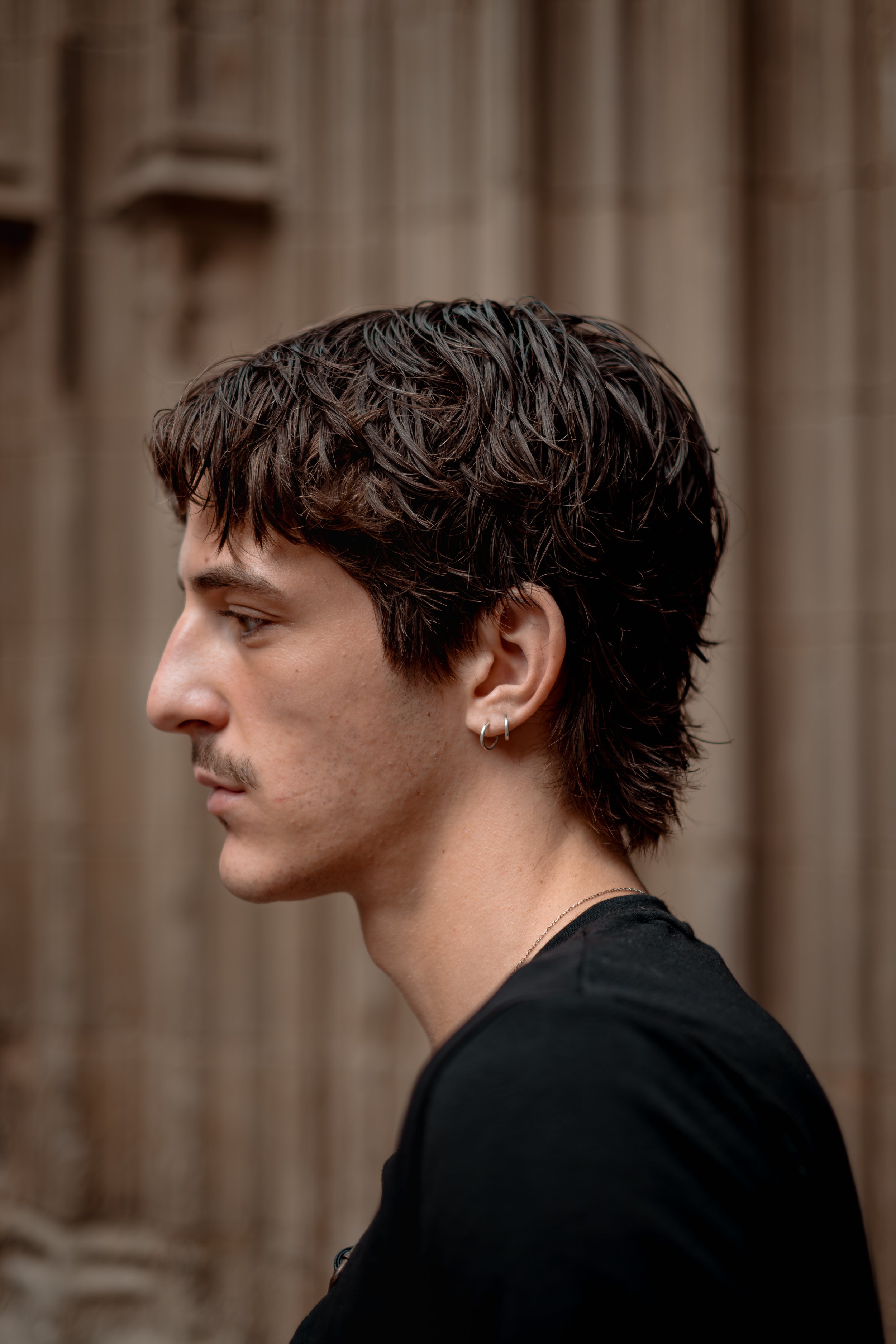 Side profile of a young man with dark, curly hair and earrings, wearing a black shirt, standing against a wooden background.