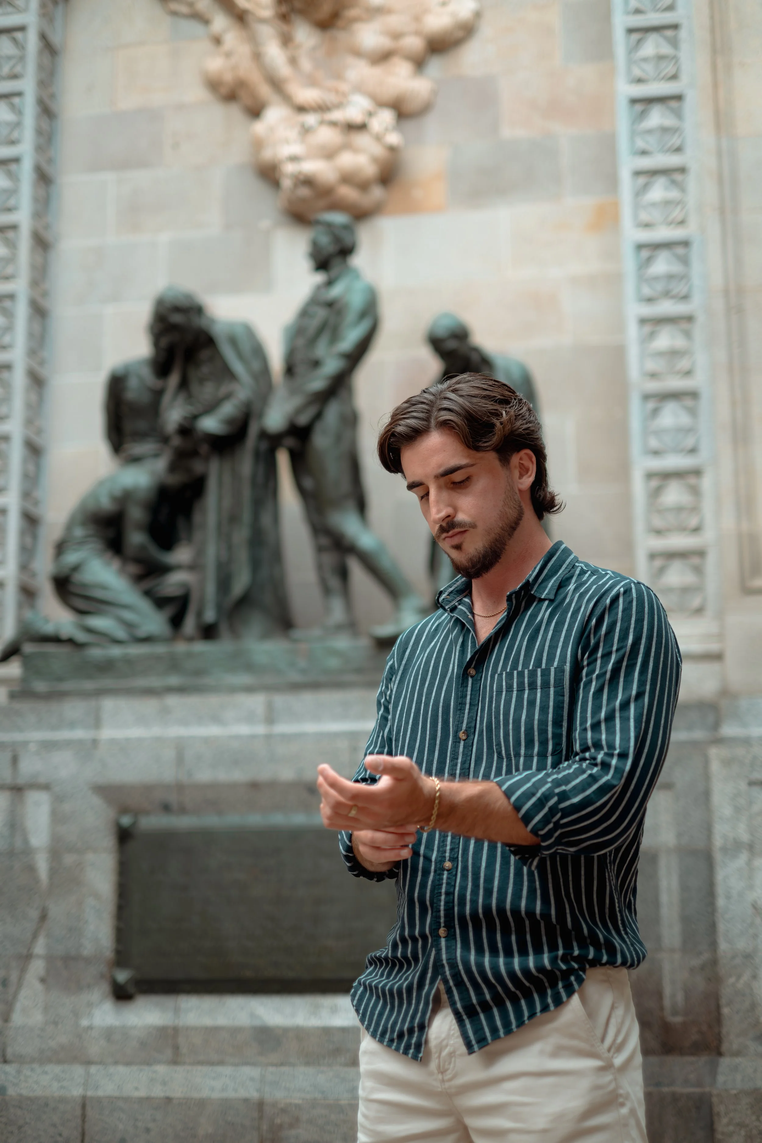 A young man with dark hair and a beard standing in front of a large bronze sculpture inside a building, looking at his phone.
