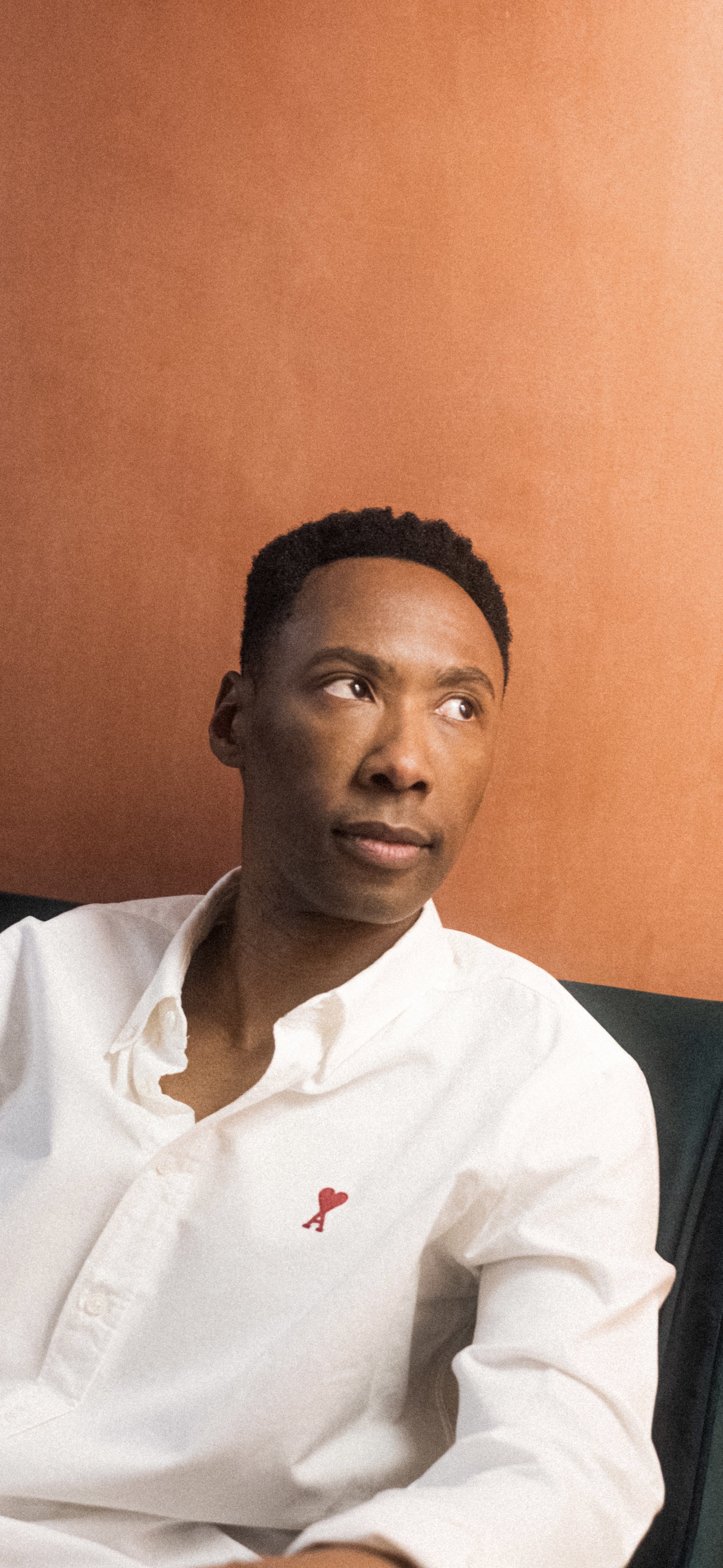 A young man with short curly hair wearing a white button-down shirt with a red heart and arrow patch on the left chest, sitting against an orange wall, looking slightly to the right.