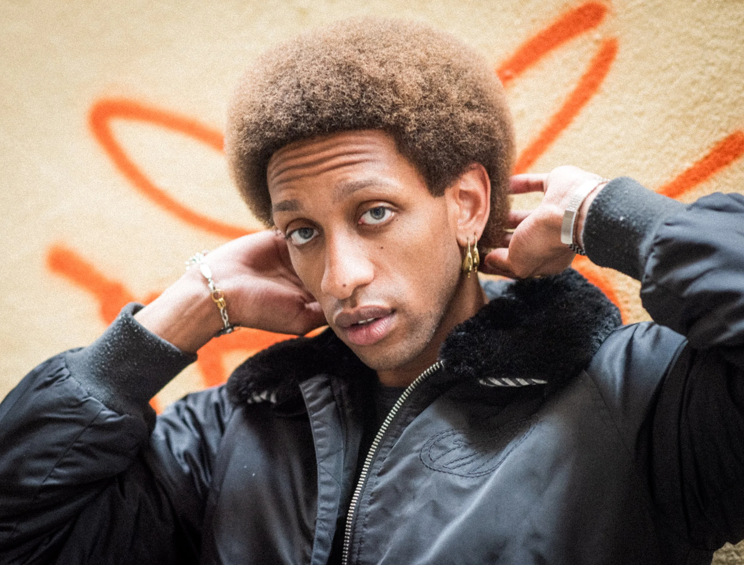 A person with brown afro hair and light blue eyes posing with hands behind their head, wearing earrings, a bracelet, and a black jacket with faux fur collar against an orange graffiti background.