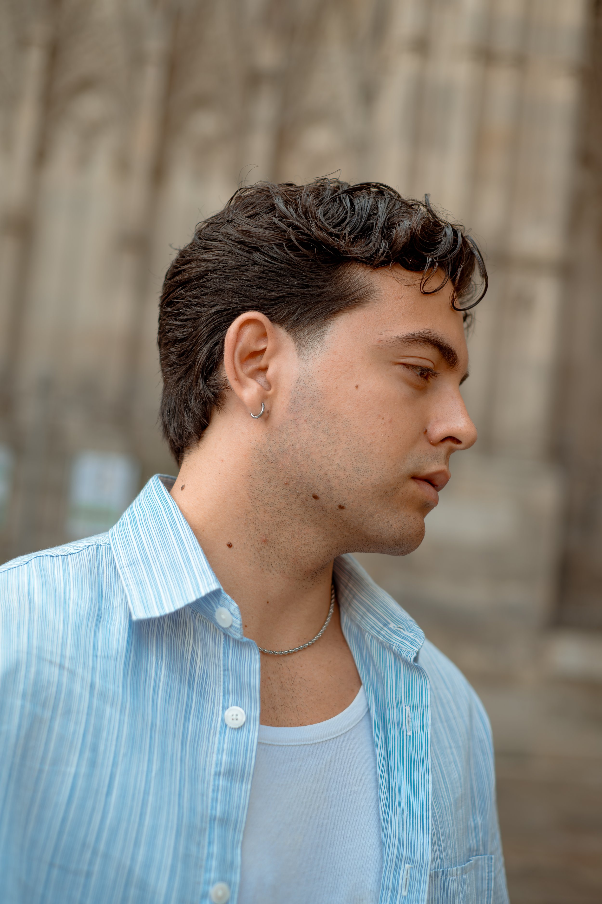 Side profile of a young man with dark, wavy hair, wearing a light blue striped shirt and a white T-shirt, with earrings and a necklace, outdoors with blurred trees in the background.
