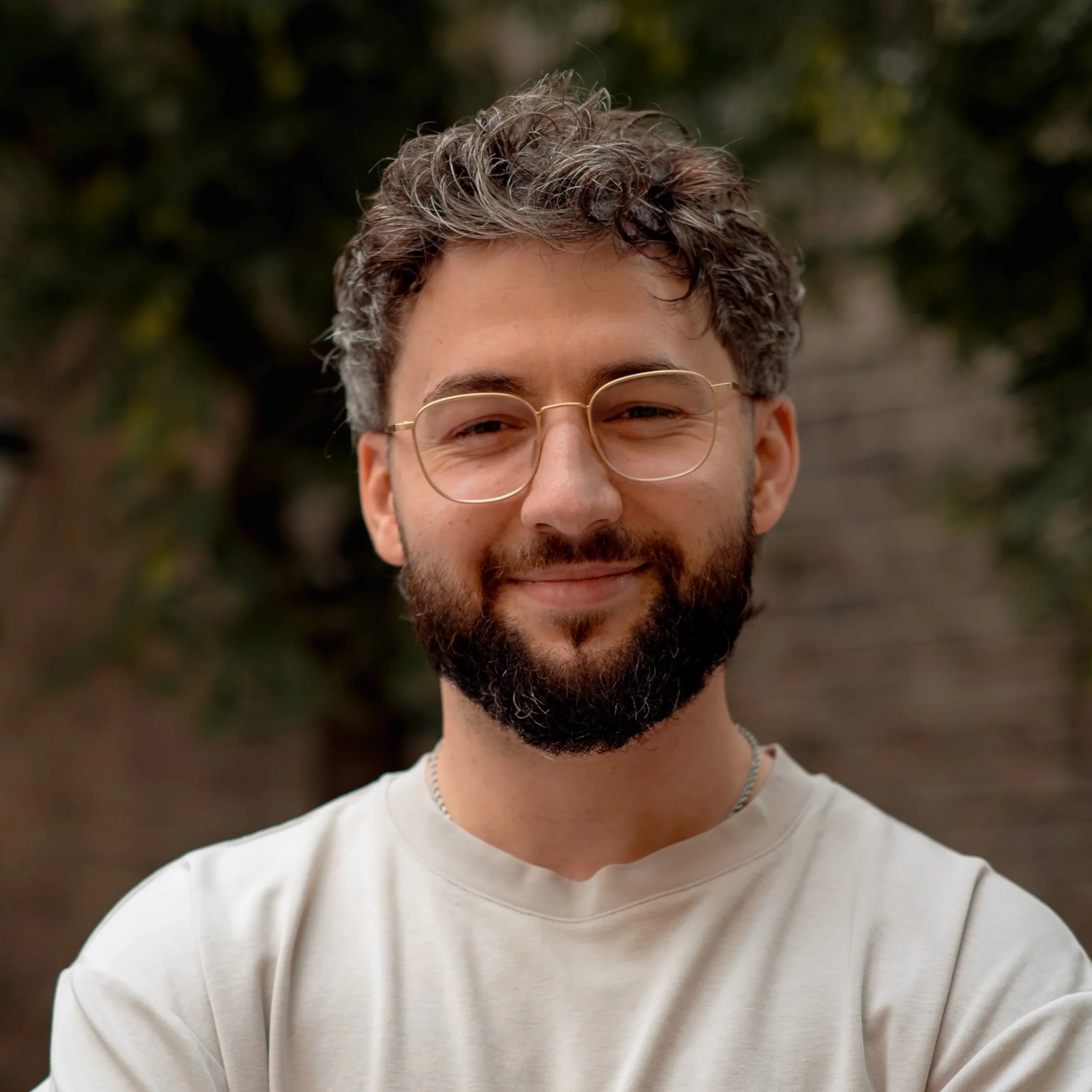 A young man with curly hair, glasses, and a beard smiling outdoors against blurred greenery.