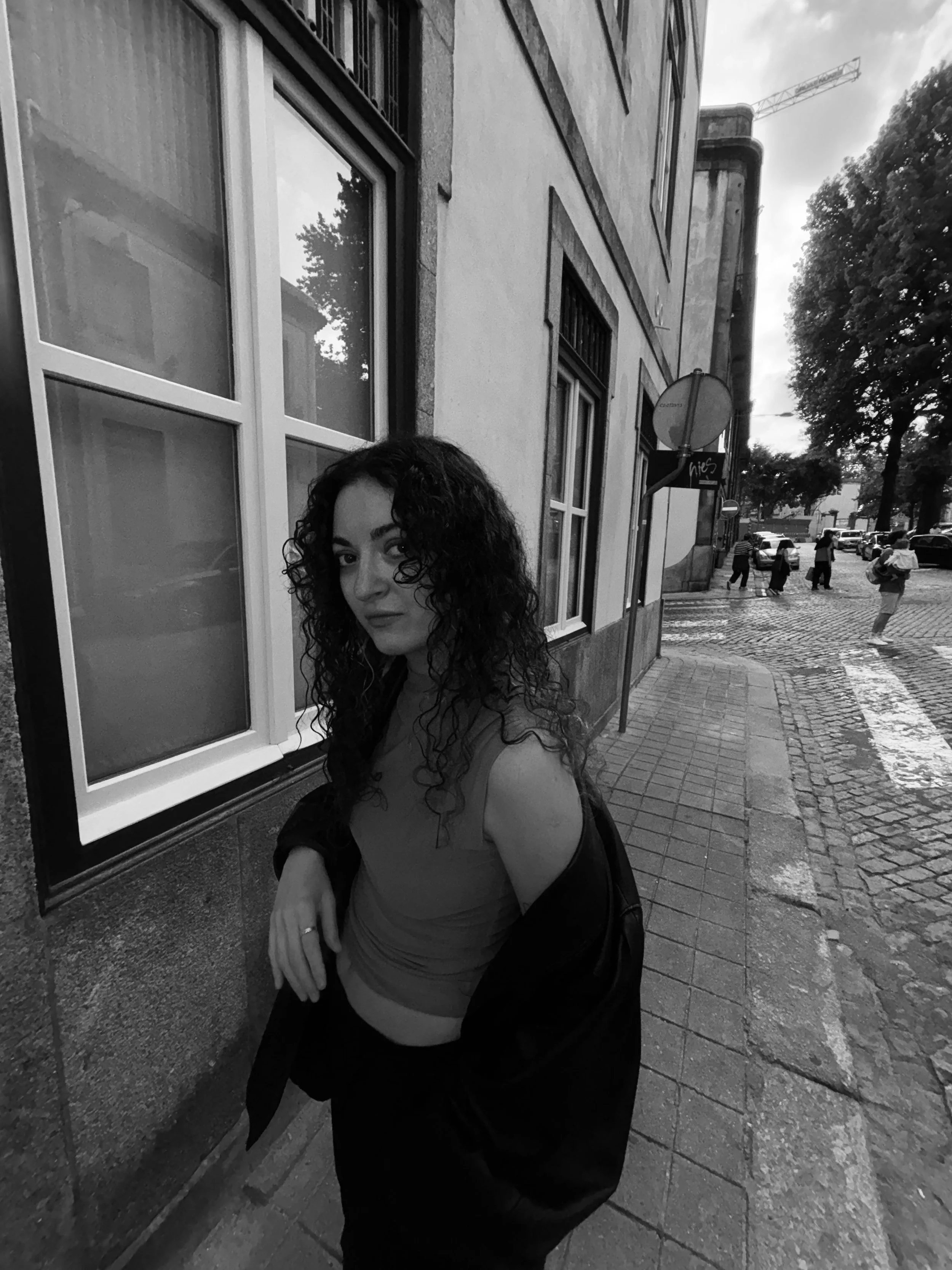 A woman with curly hair leans against a building on a city sidewalk, looking at the camera, in black-and-white photo.