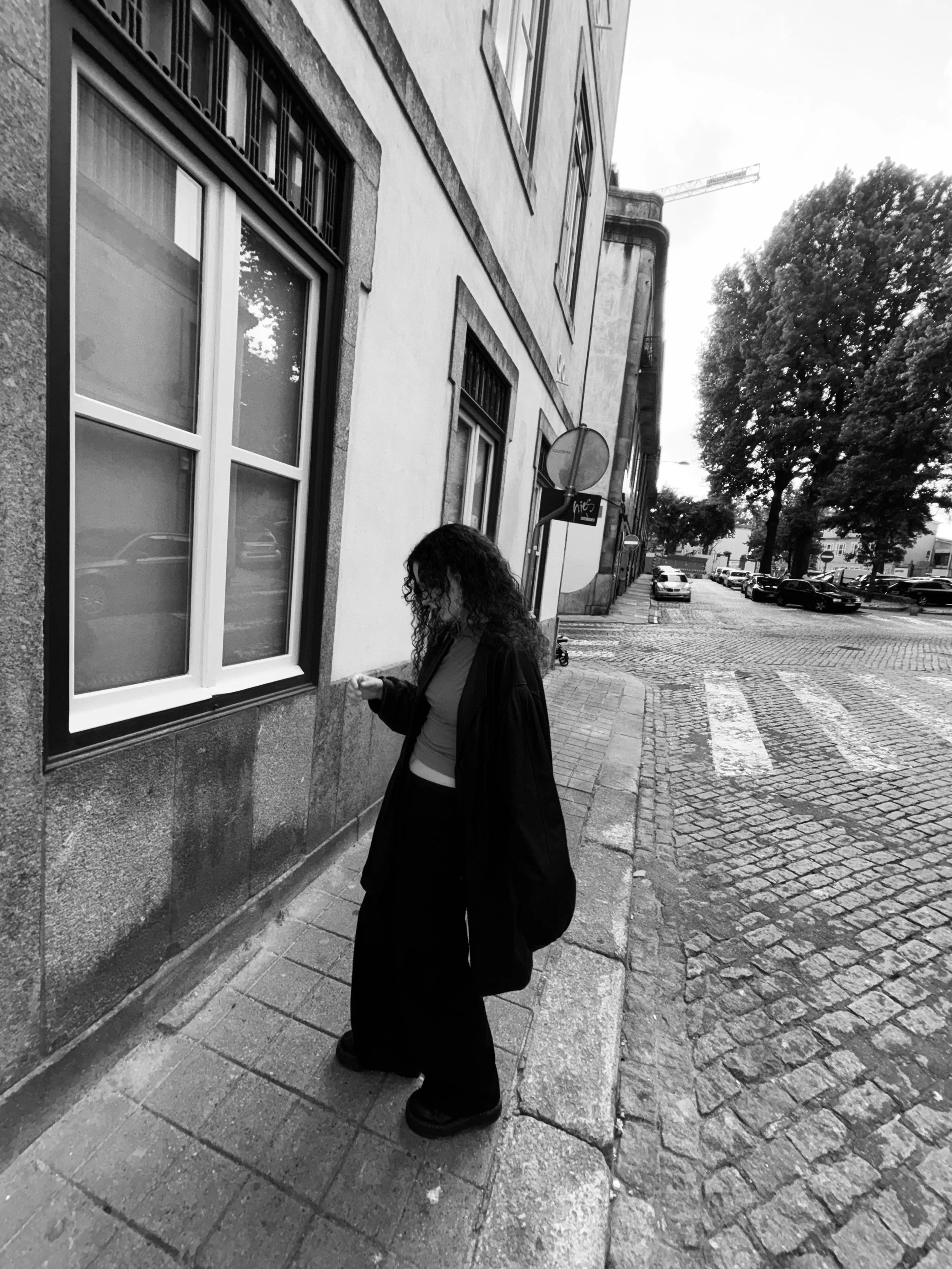 A woman with curly hair and glasses standing on a cobblestone sidewalk, looking at her phone, next to a building with large windows, in a city street scene in black and white.