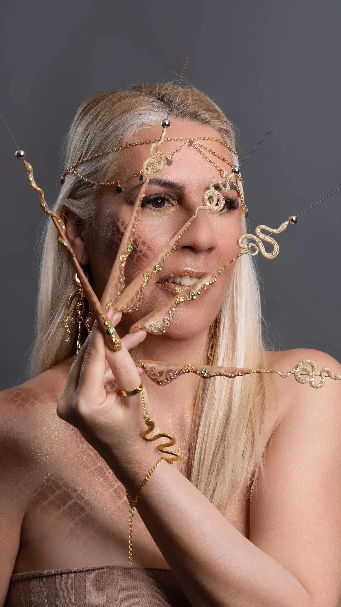 A woman with long blonde hair and gold jewelry covering her face, holding a gold chain with snake and circle designs against a gray background.
