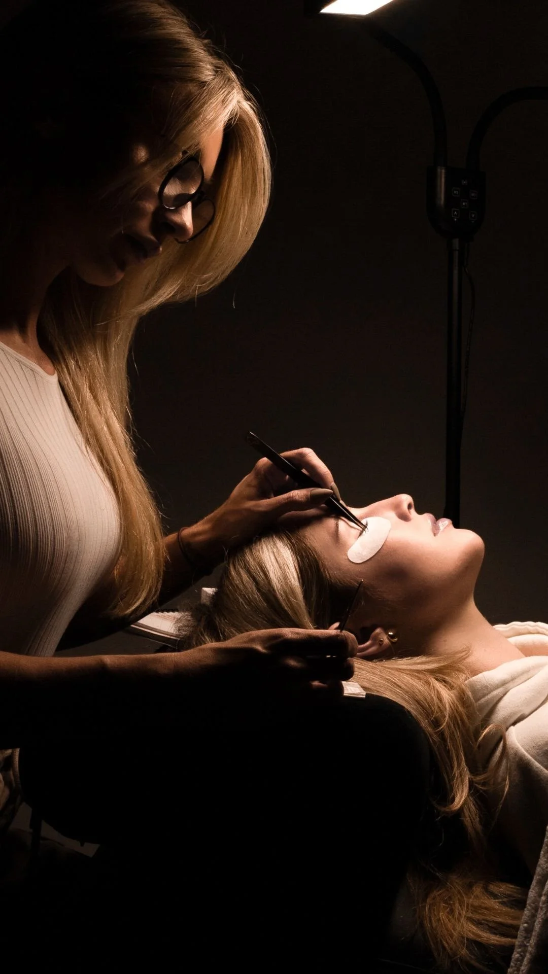 A woman lying down with her eyes closed, receiving a cosmetic eyelash extension treatment from a technician.