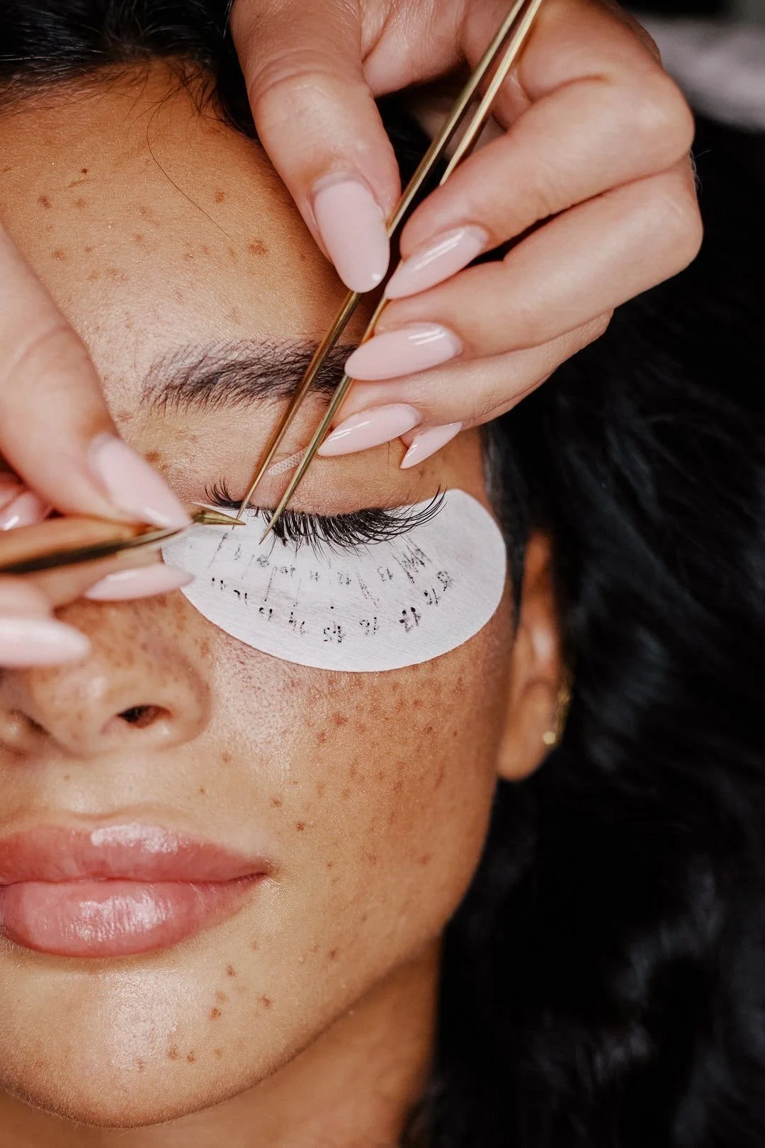 A woman is getting her eyelash extensions applied by a technician who is using tweezers. The woman is lying down with a protective pad under her eye, and her face shows freckles and smooth skin.