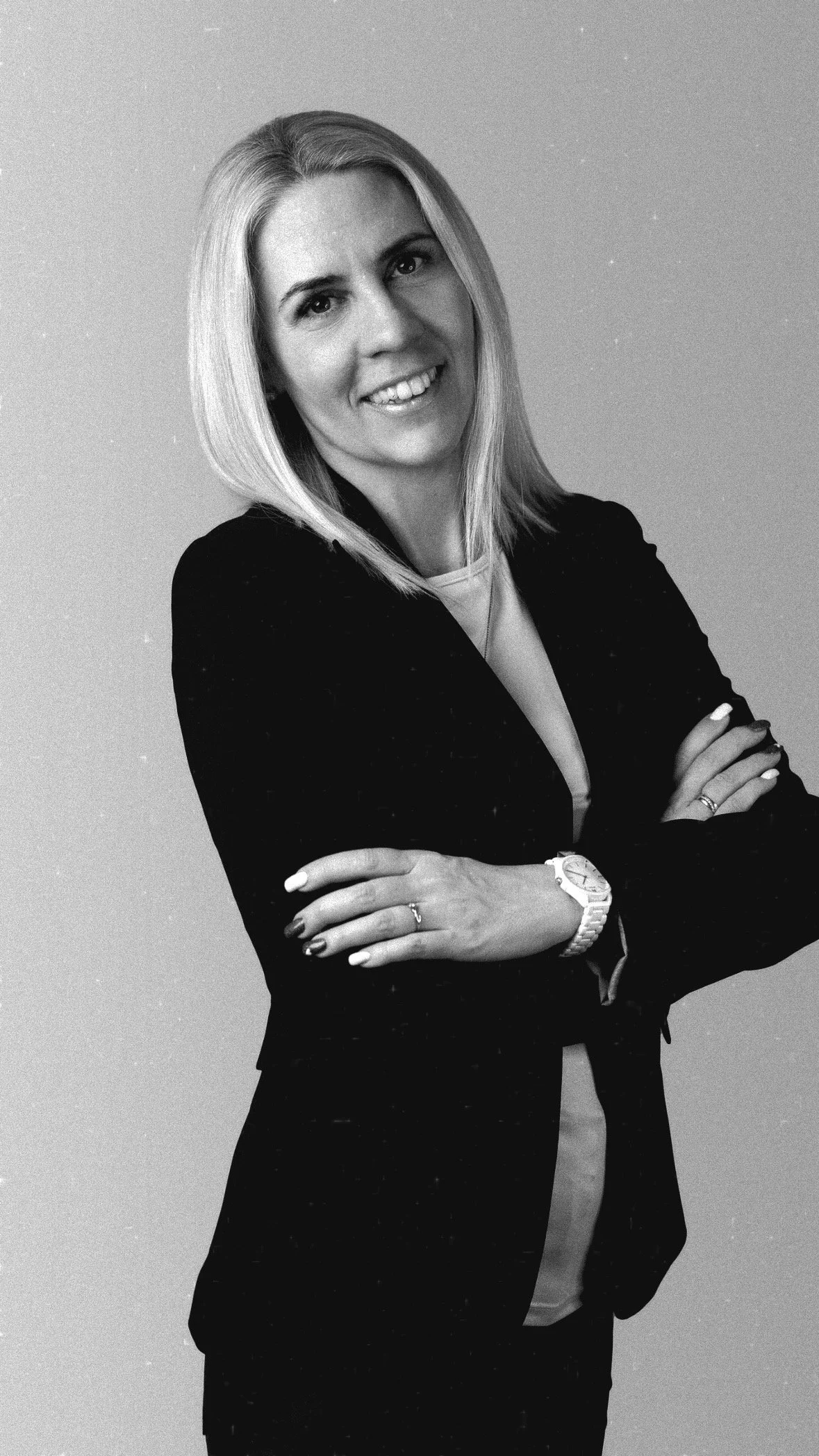 Black and white photo of a professional woman with shoulder-length blonde hair, wearing a dark blazer, white top, with her arms crossed, smiling.