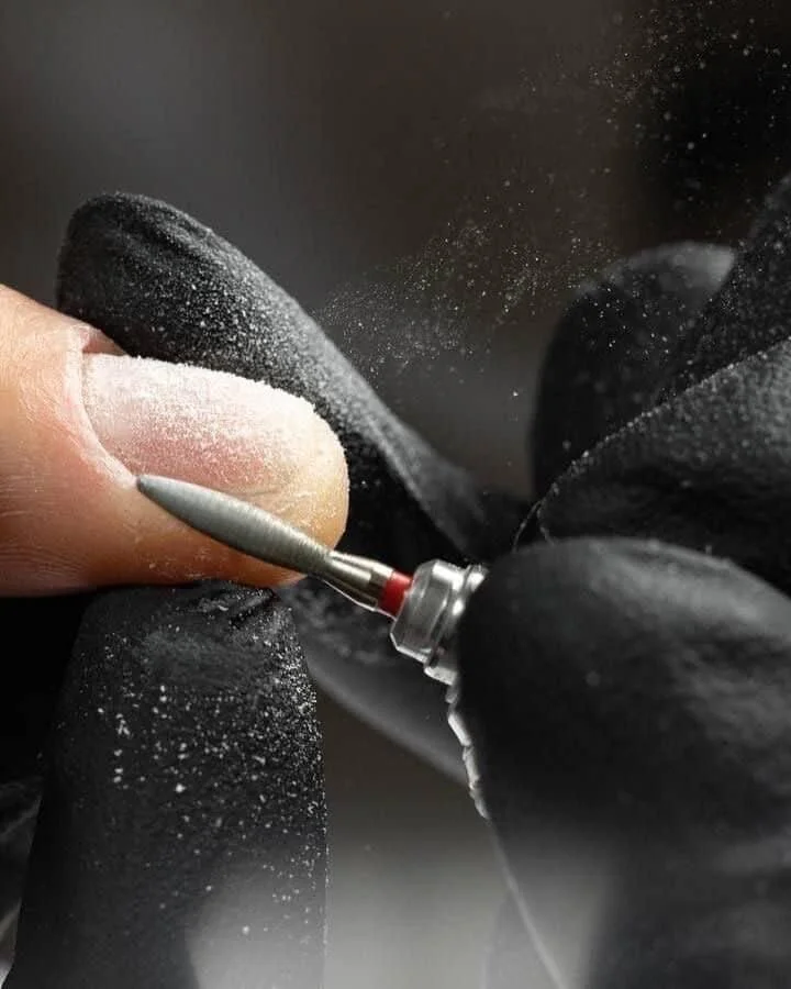Close-up of a person's finger holding a small drill, wearing black gloves, drilling into the false nail of their fingernail, creating dust from the nail.