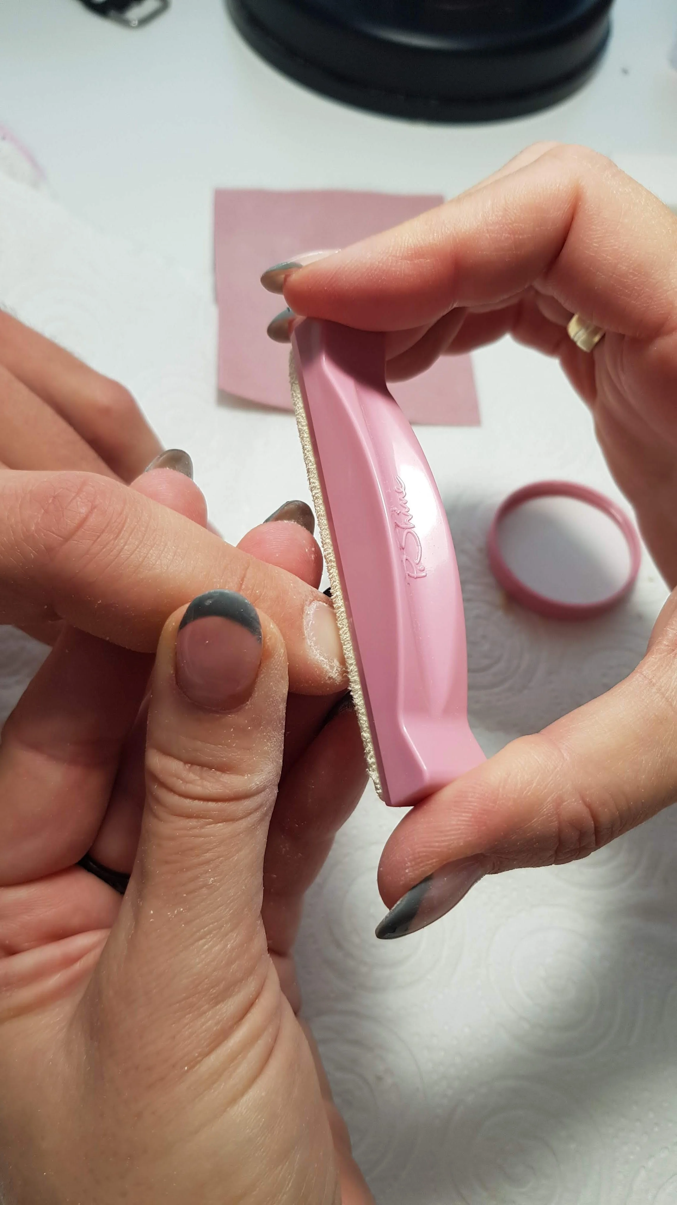 A person using a pink emery board to smooth their toenails with a white towel and pink paper in the background.