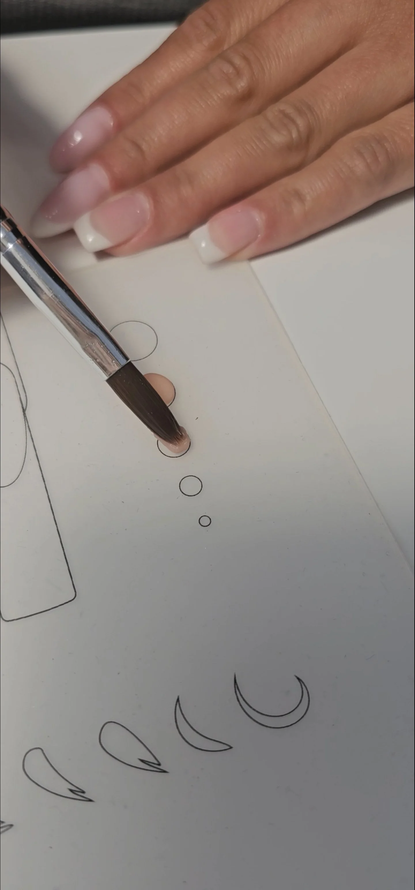 Close-up of a hand with manicured nails holding a black pen, drawing moon shapes and circles on a white paper.