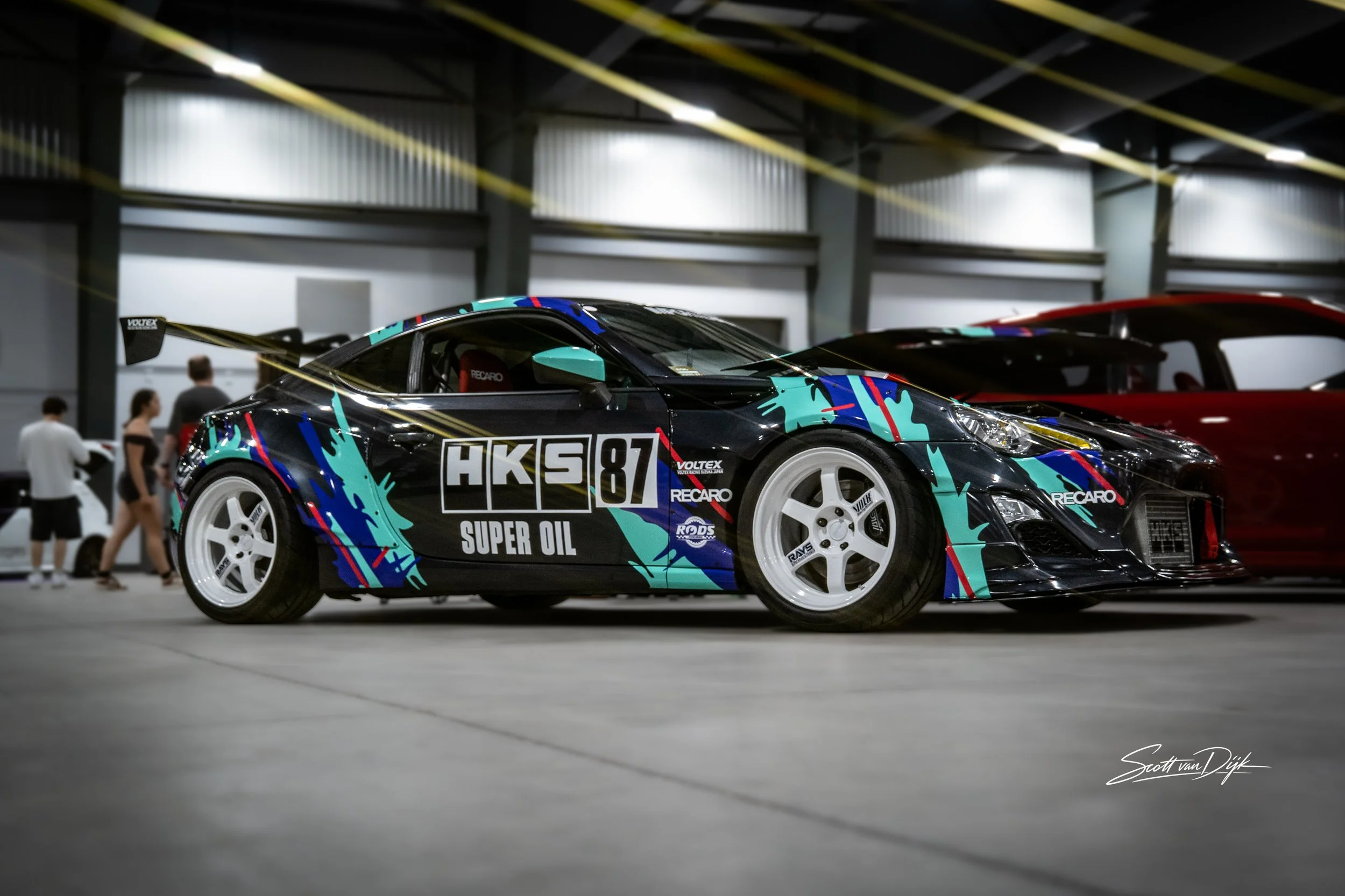 A black race car with colorful blue, red, and yellow splash patterns and the words 'HKS 87 Super Oil' on the side. It is displayed indoors at what appears to be an exhibition, with a high ceiling and metallic walls. People are walking around in the background.
