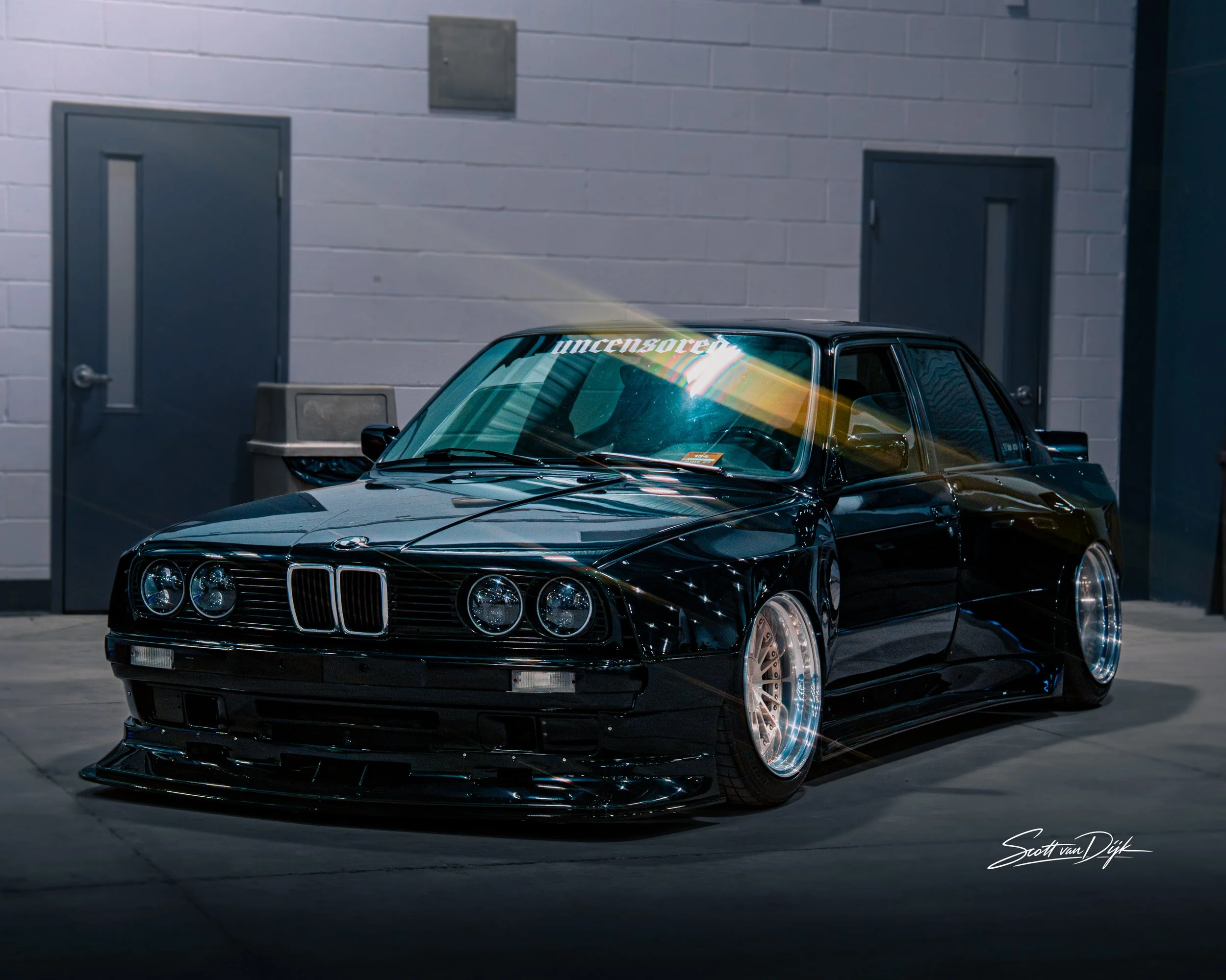 A black BMW E30 car parked in an indoor parking garage with gray brick walls and gray doors.