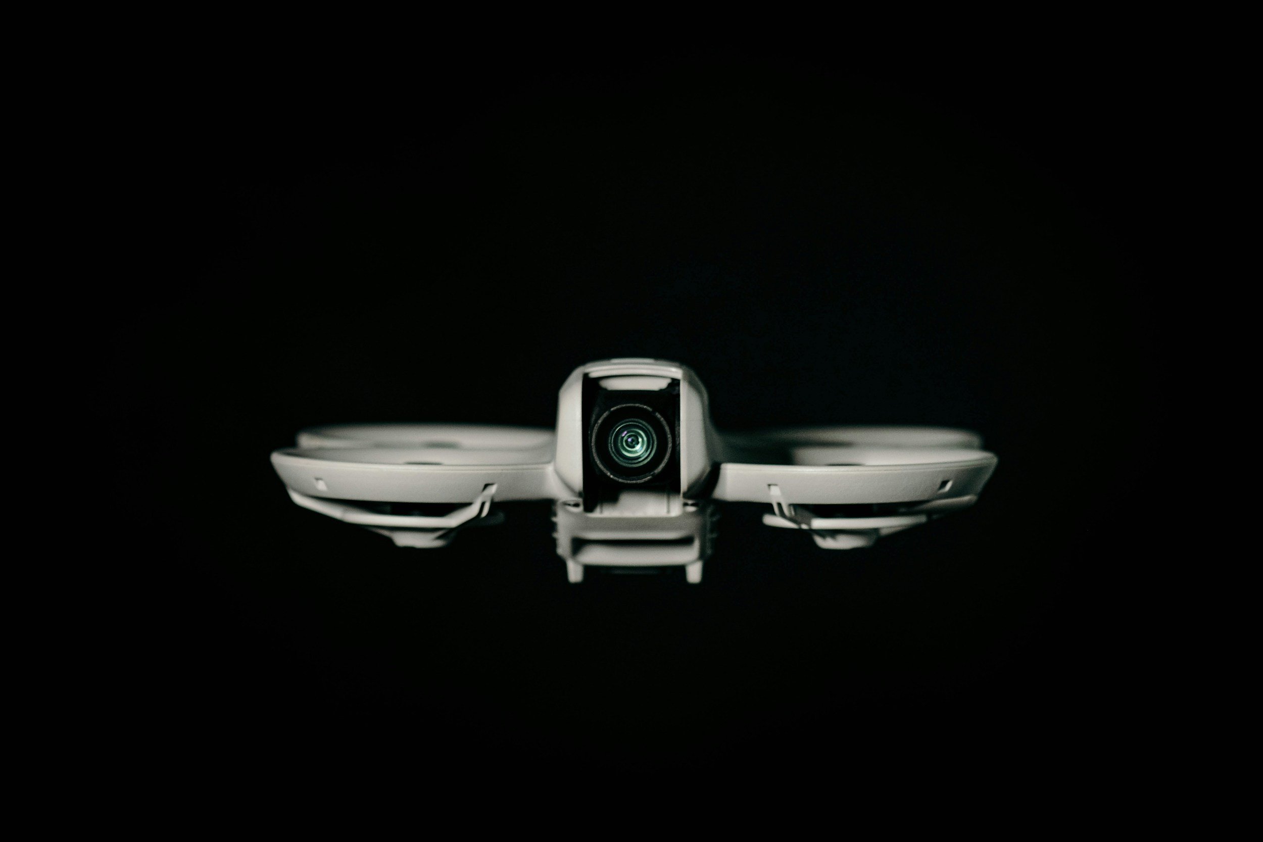 A white drone with a camera lens on the front, viewed from below, set against a black background.