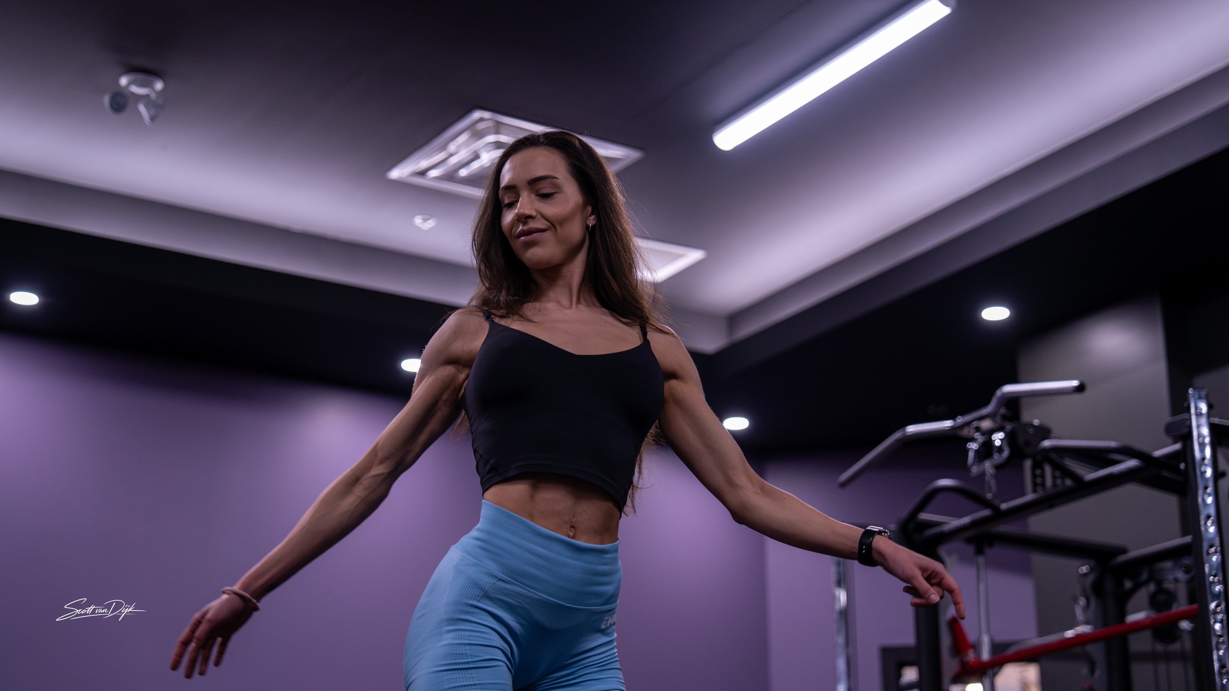 A woman fitness trainer with tan skin and long brown hair exercising in a gym, standing with arms extended and smiling. Body building, bikini posing.
