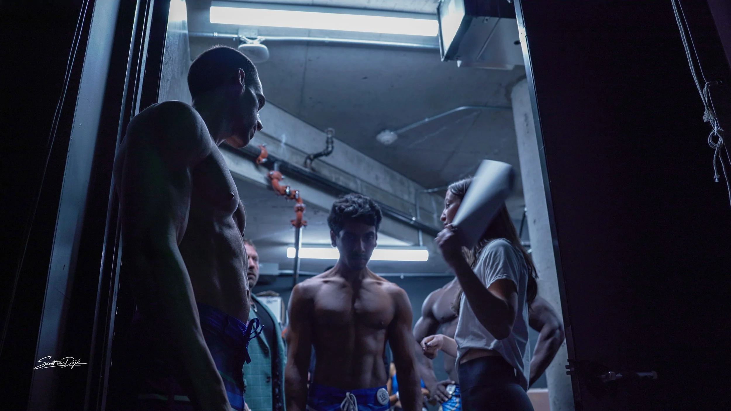 A group of people in an industrial setting, with two shirtless men in the foreground, one looking down, and one woman holding a clipboard, possibly preparing for or participating in a fitness or training session. Body building Ottawa.