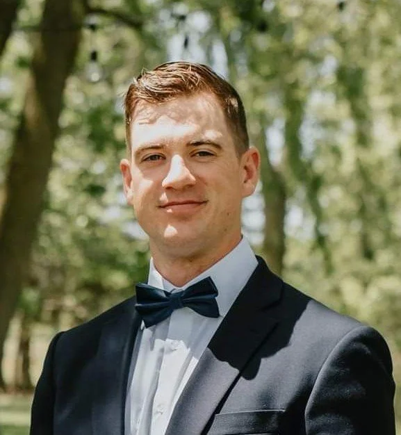 Young man in a tuxedo with a bow tie outdoors, smiling, with trees in the background.