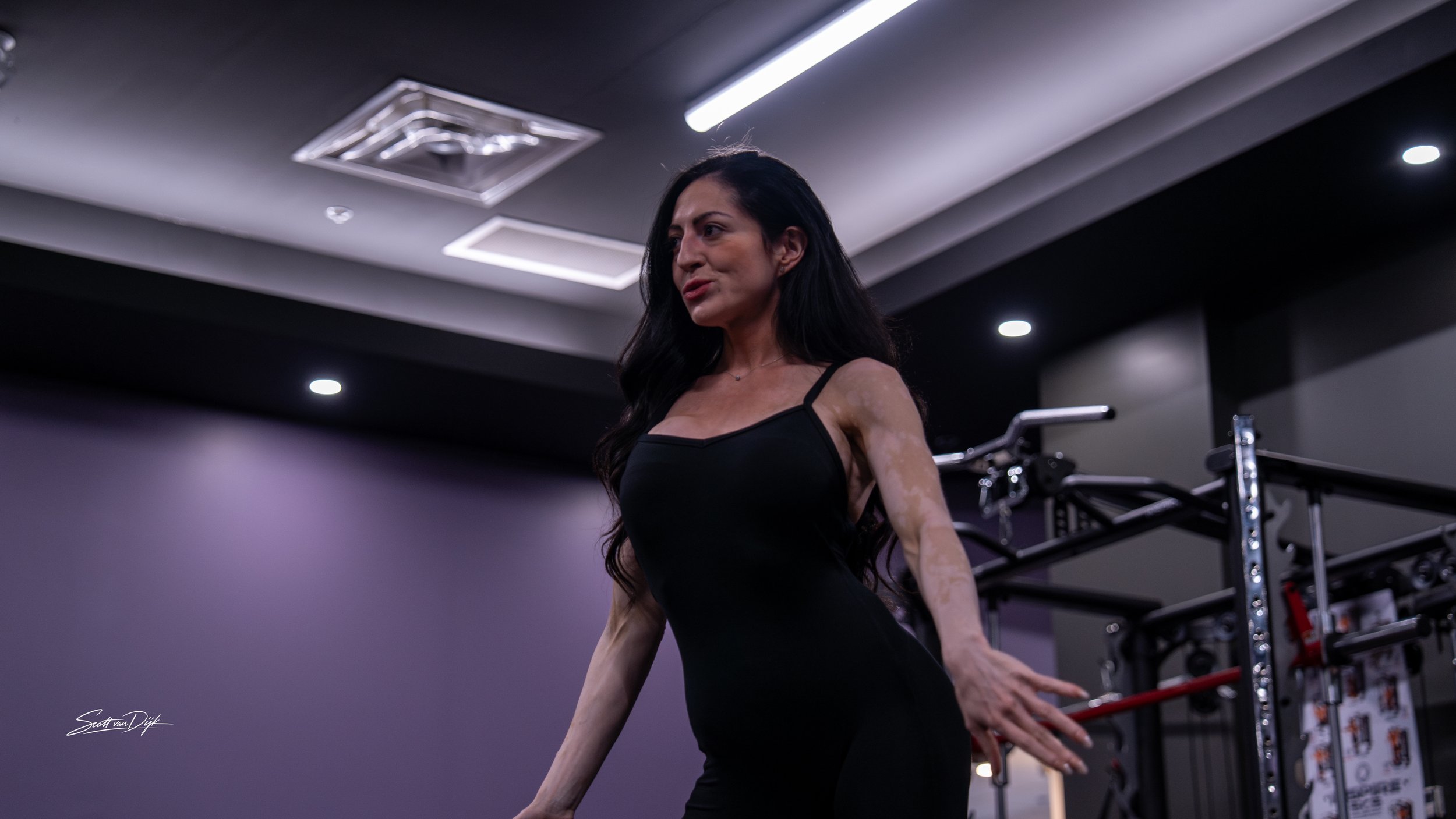 Woman with long black hair in a black dress in a gym or fitness studio, standing next to workout equipment, with ceiling lights and dark-colored walls in the background. Fit model body building posing coach.