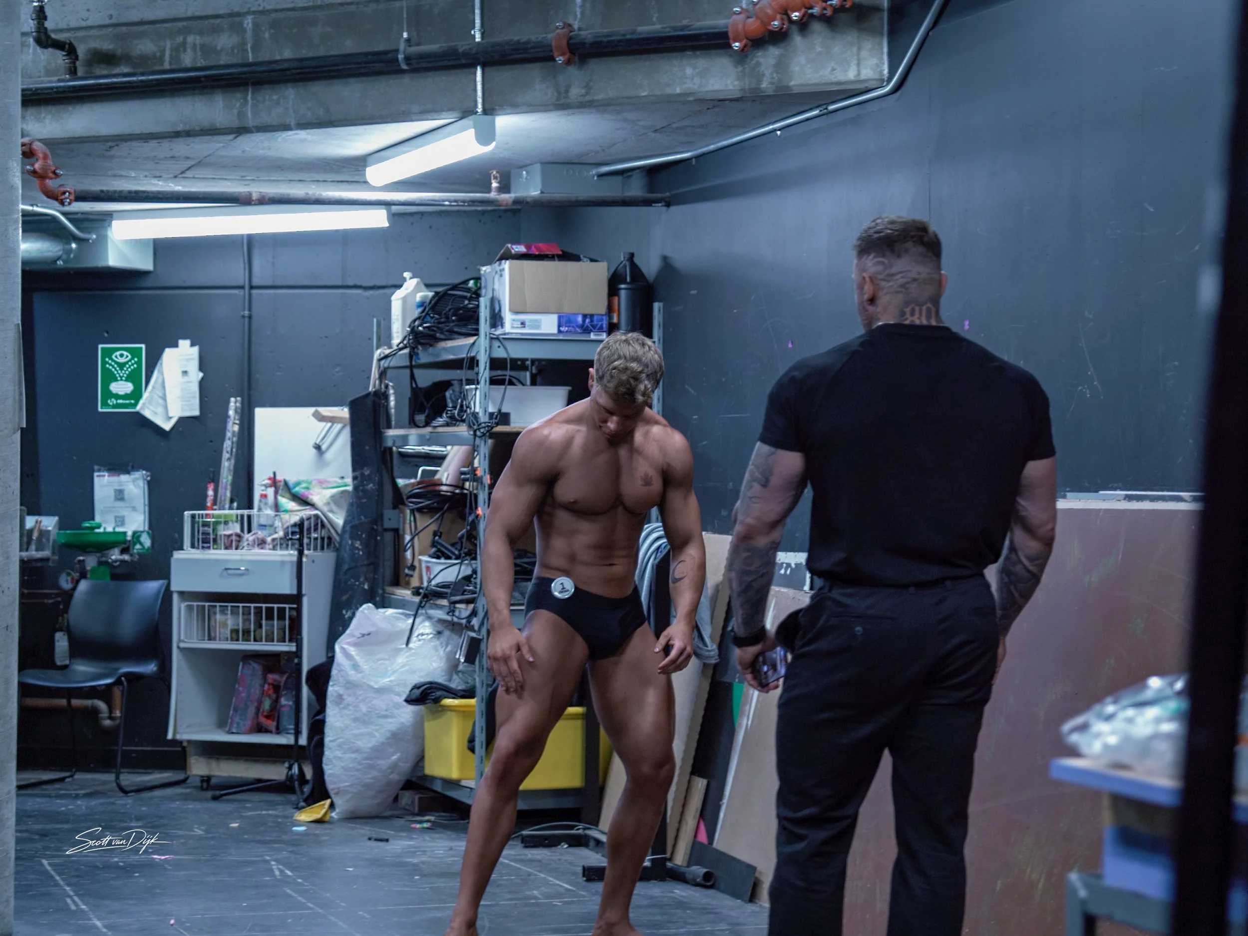 A topless man with tattoos on his chest and abs is standing in an indoor, industrial-style space, being assisted by another man dressed in black. Backstage Body building photography.