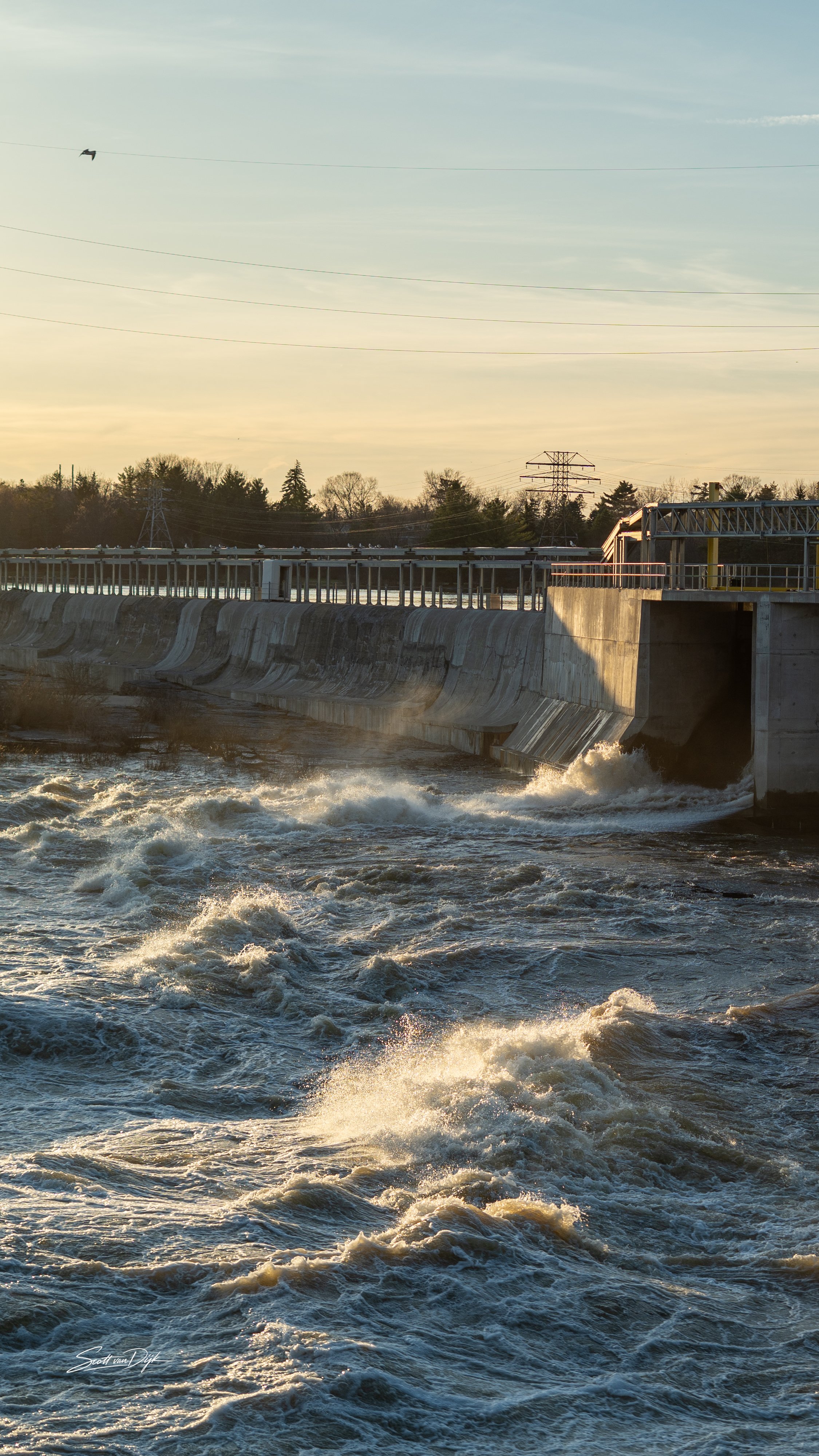 A dam with water flowing through it, an electrical tower in the background, and a partly cloudy sky with a faint sun. Ottawa Canada
