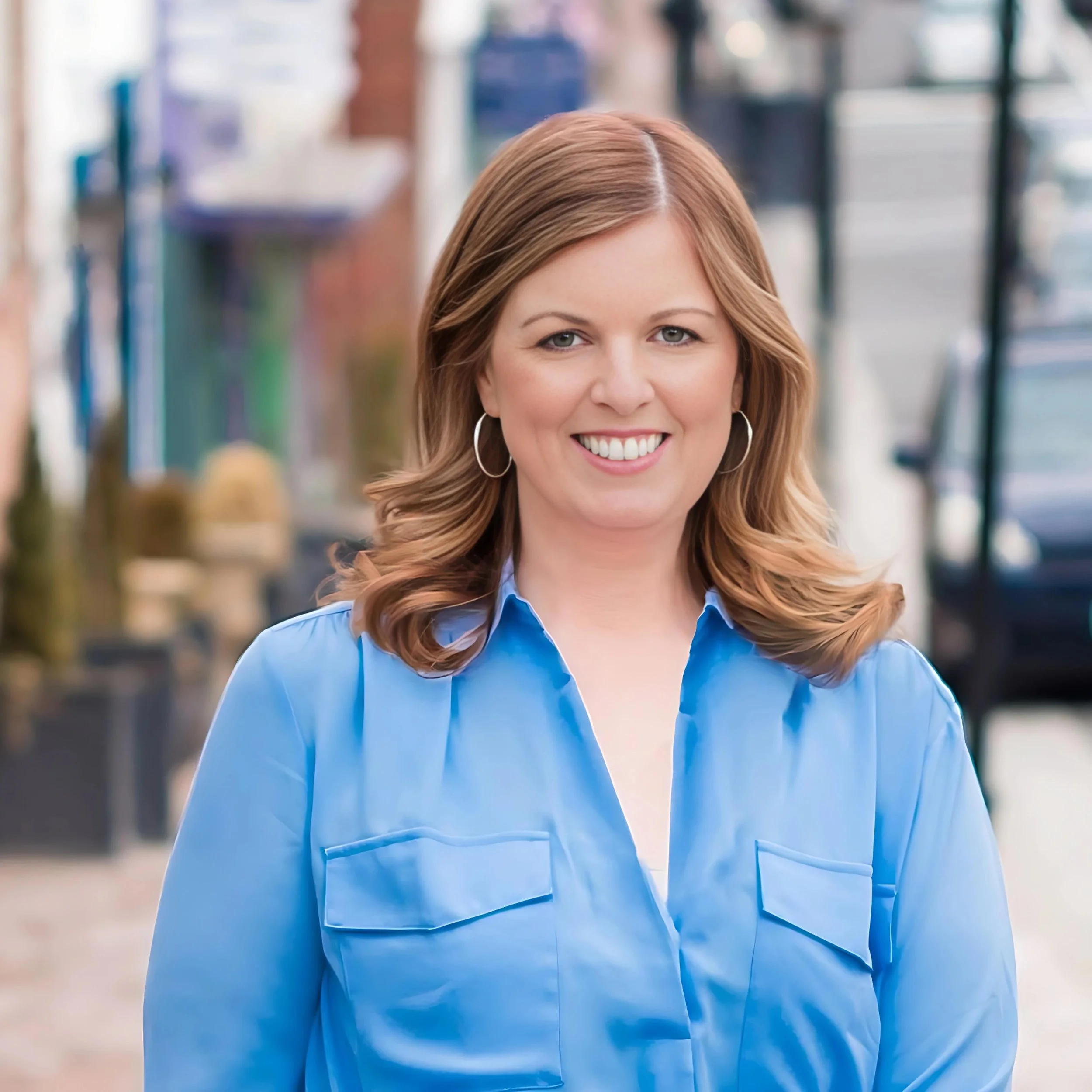 A smiling woman with shoulder-length wavy hair wearing hoop earrings and a blue button-up shirt, standing outdoors in an urban setting.
