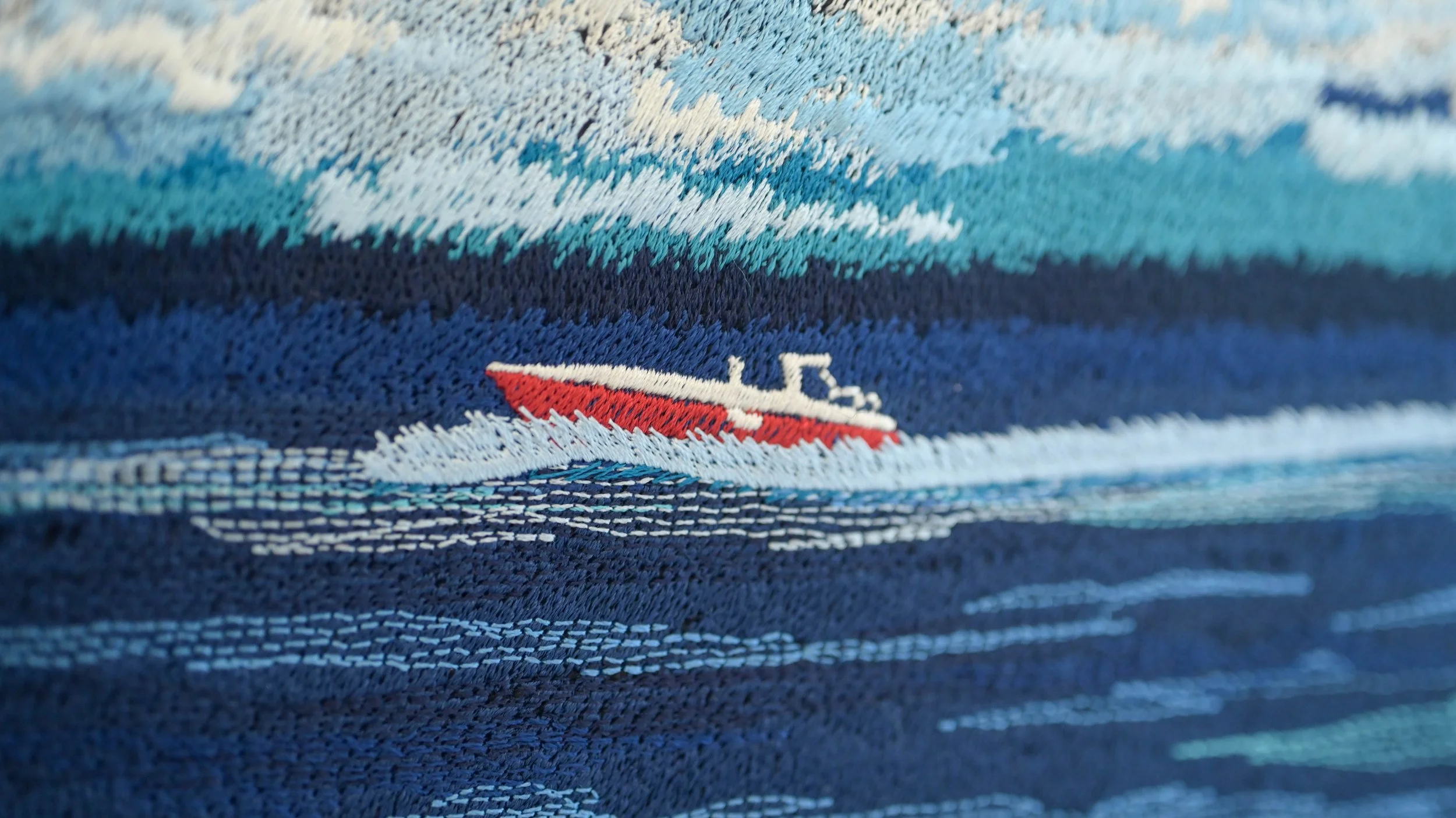 Close-up of a blue, white, and red embroidered fabric depicting a boat on water, with waves and sky in the background.