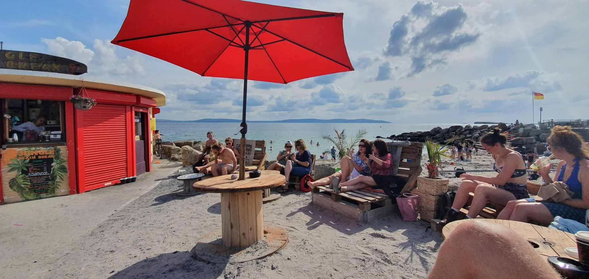 Beach scene with people sitting on wooden benches and relaxing, a red umbrella providing shade, a small red beach bar, and the ocean in the background with a cloudy sky.
