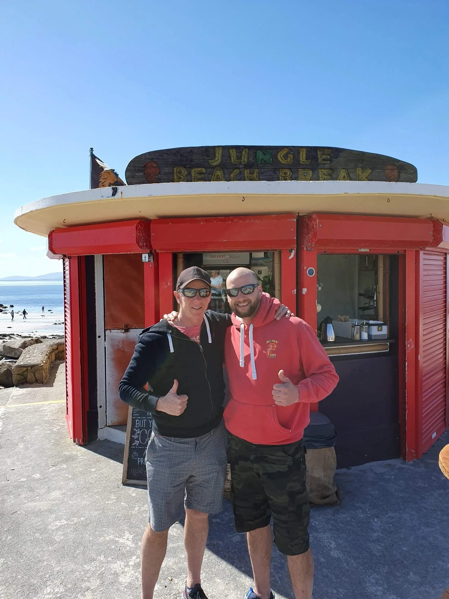Two men standing together, giving thumbs up in front of a red beachside kiosk with a sign that reads "Jungle Beach Break." The background shows a beach with people, the ocean, and a clear blue sky.