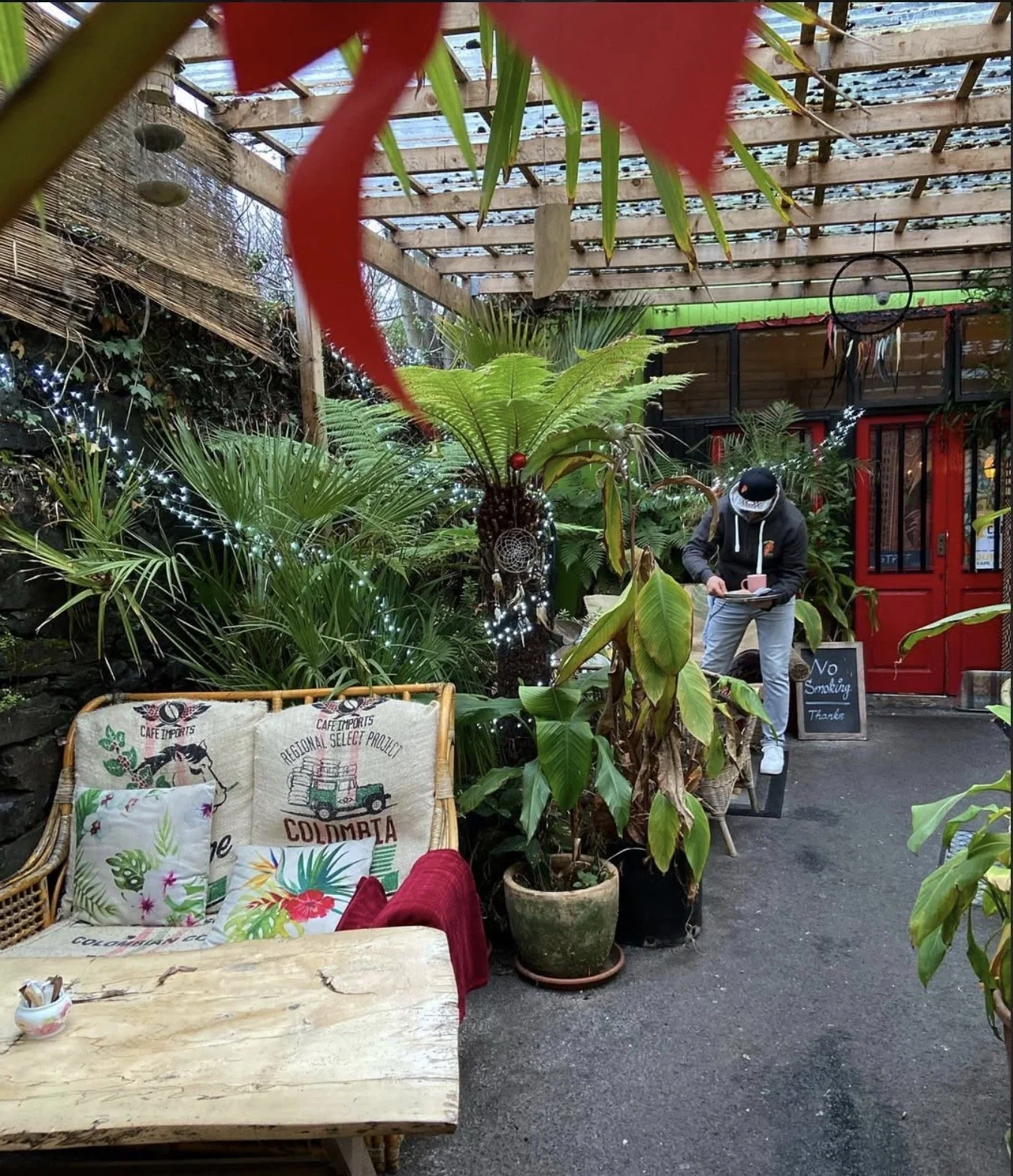 Cozy outdoor cafe with lush green plants, a wooden table, patterned cushions, fairy lights, and a person reading a menu near a red door with a 'No Smoking' sign.