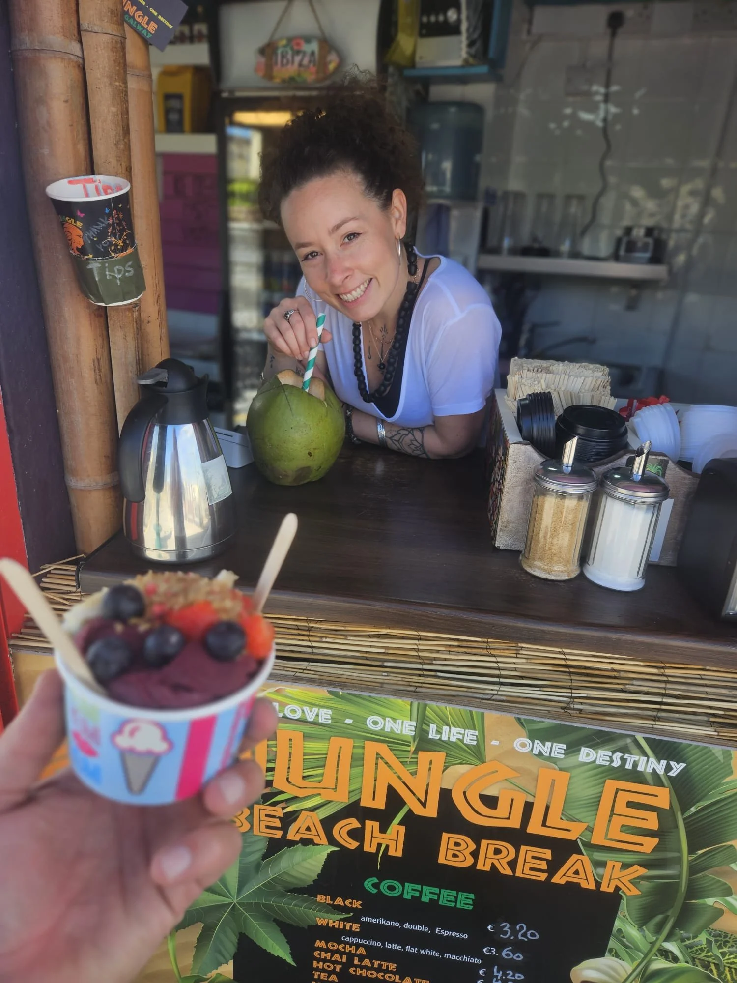 A woman smiling at a beachside kiosk with a coconut drink. The kiosk offers various coffee beverages listed on a menu. The woman is wearing a white shirt, has tattoos, and is holding a green coconut with a straw. In the foreground, a hand holds a cup of berry ice cream with blueberries, strawberries, and other berries.