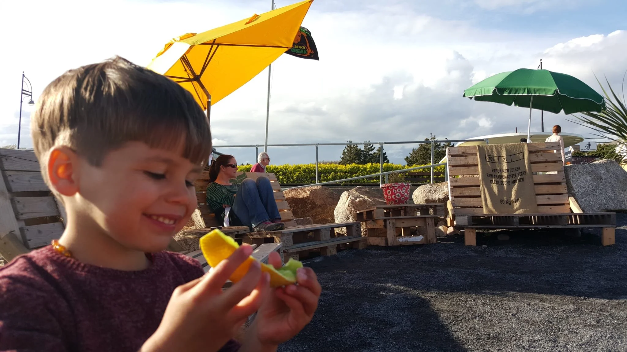 A young boy with brown hair smiling while holding a slice of lemon and a lime on a sunny outdoor patio with several people sitting on wooden benches under green and yellow umbrellas.