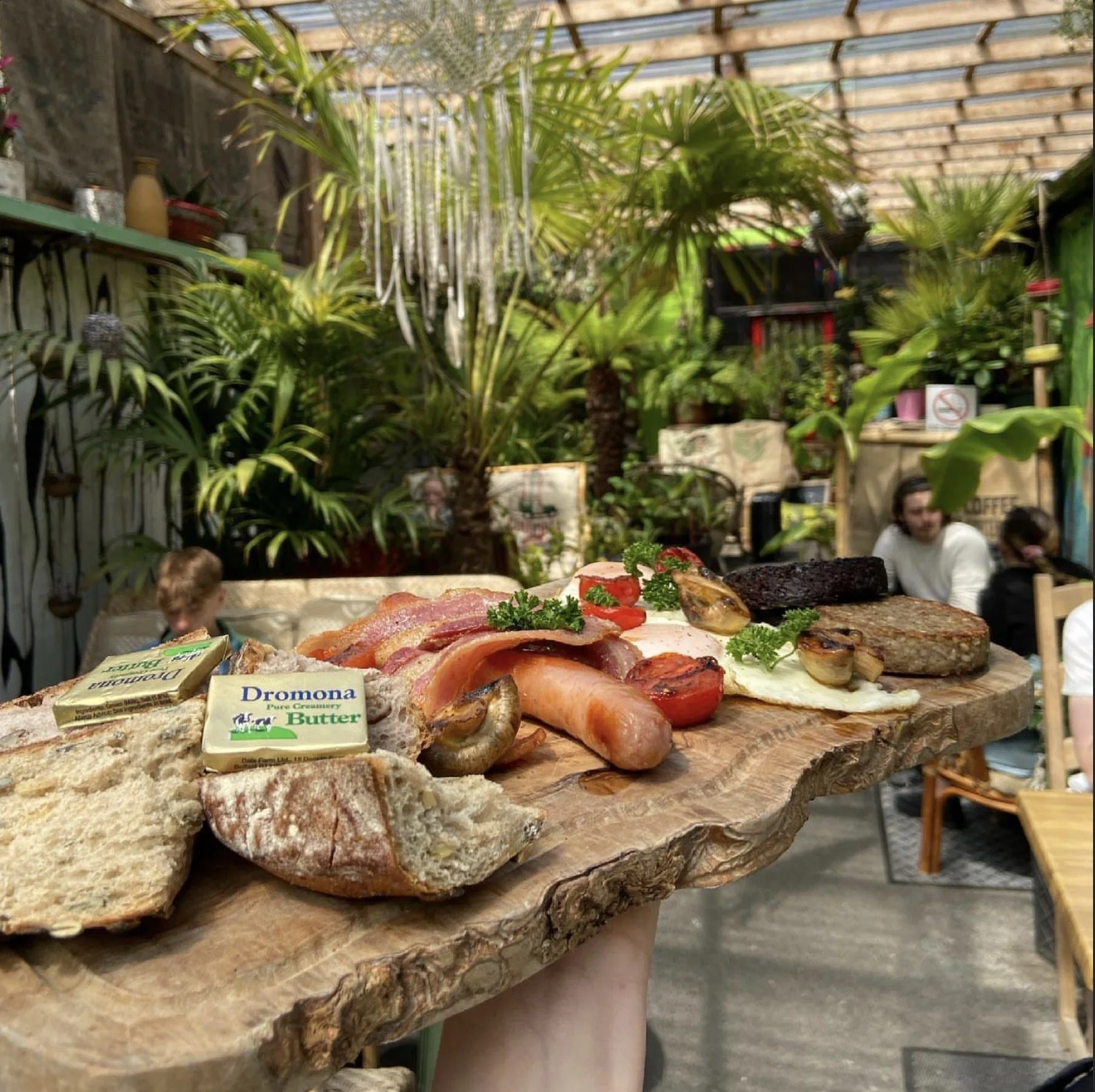 A wooden platter with various breakfast foods including slices of bread, bacon, grilled vegetables, herbs, and sausage. In the background, there are lush green potted plants and a cozy dining area with people sitting at tables in a greenhouse or garden setting.
